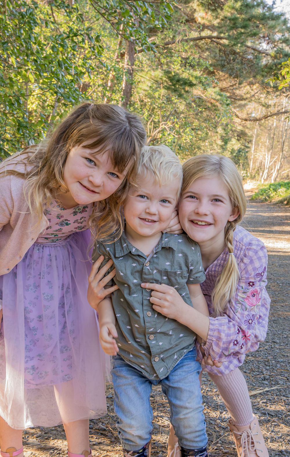 Three happy children pose together outdoors on a sunny path surrounded by green trees.