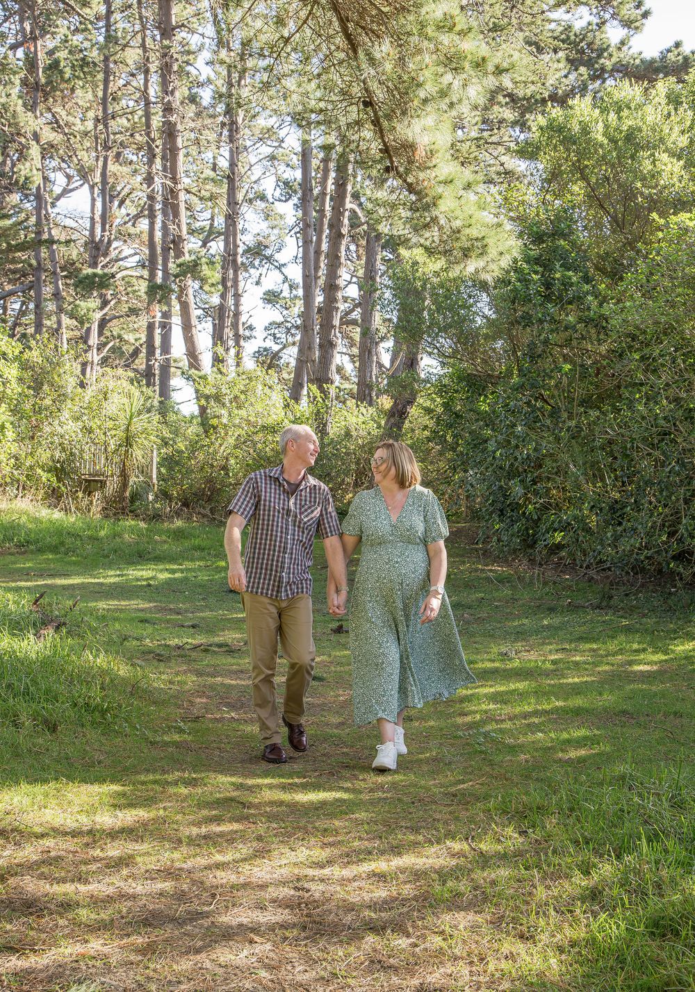 Couple holding hands walking on a sunny forest path surrounded by lush green trees and grass.