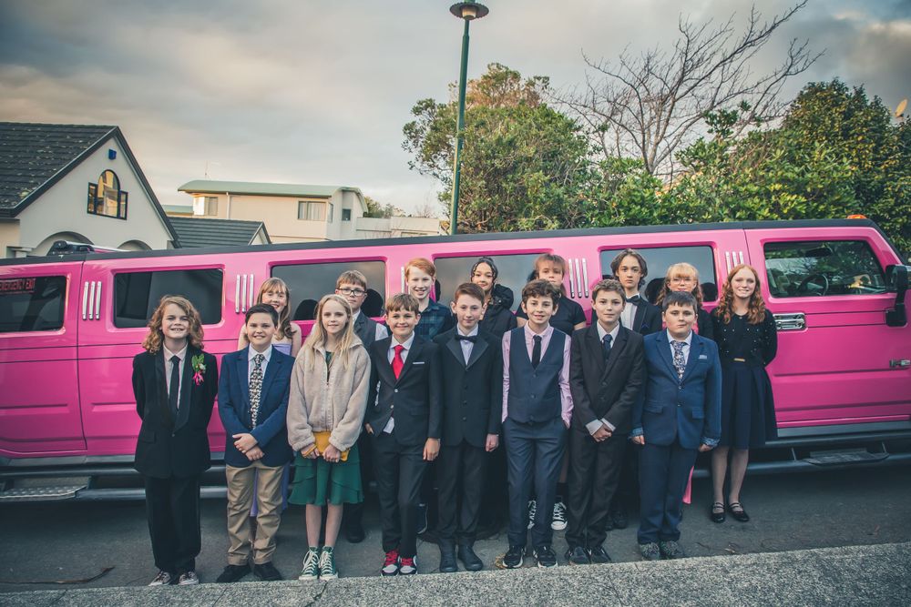 Students in formal wear line up for photo beside pink stretch limousine.