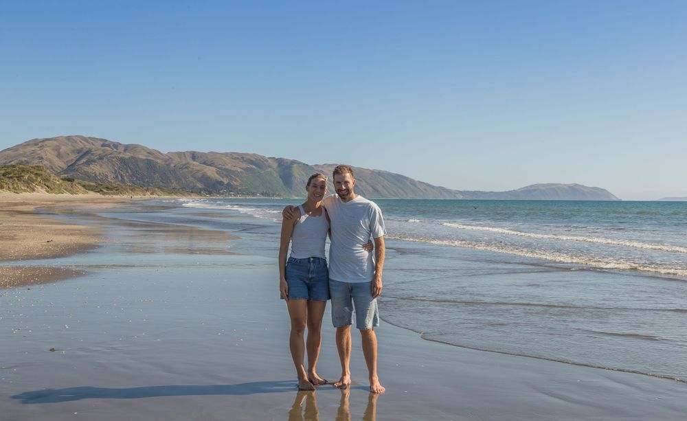 Couple standing together on wet sand with mountains and ocean creating a scenic backdrop.