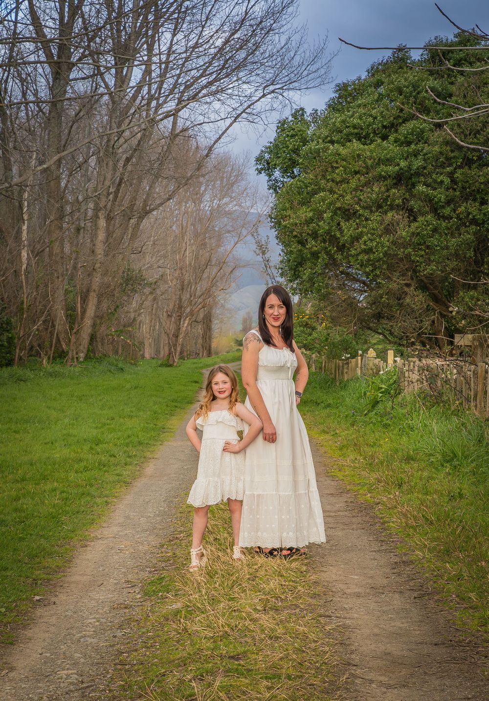A dirt path through trees and grass shows two people in white dresses walking together on a sunny spring day.