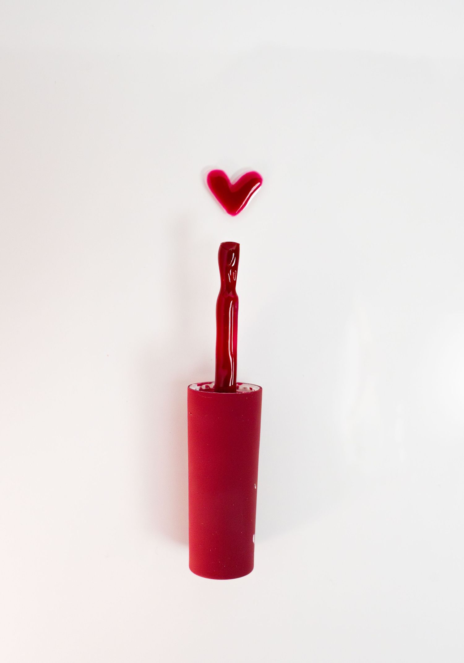 Red nail polish bottle with heart-shaped design floating above against white background.