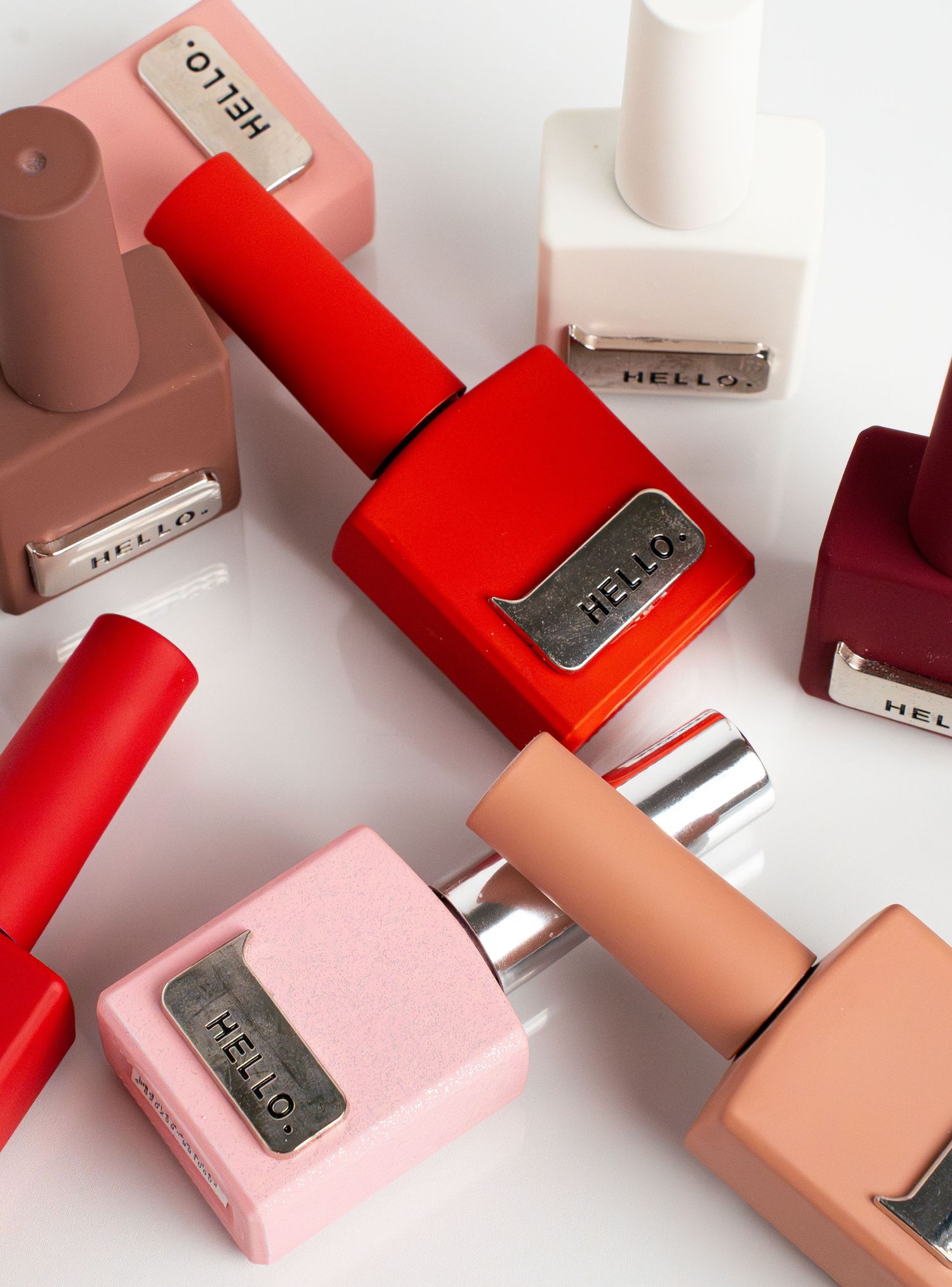 Modern nail polish bottles in shades of red, pink, and nude with metallic Hello labels.