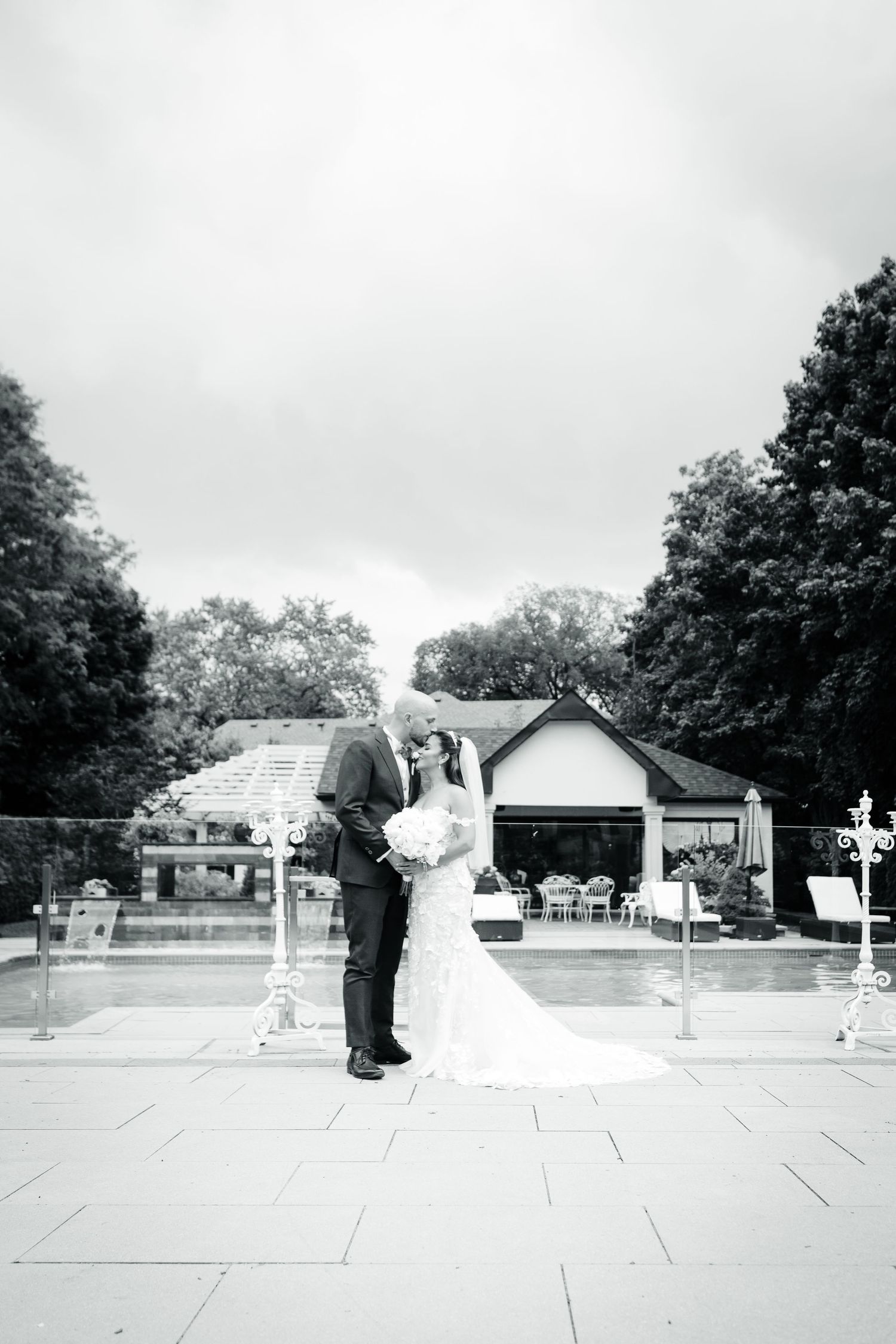 Black and white wedding photo of a couple standing by a pool with trees in the background.