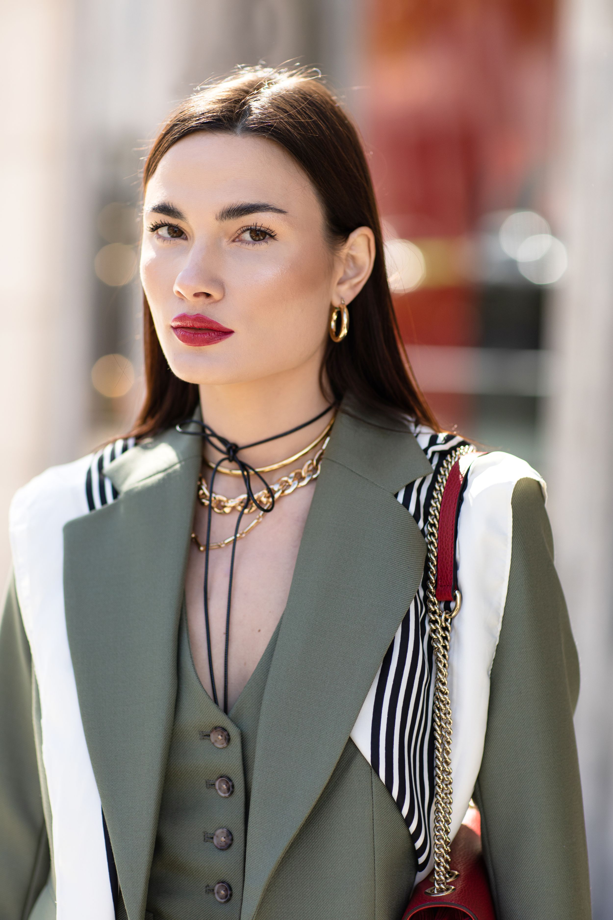 Professional fashion portrait of a model wearing a sage green blazer with layered necklaces and bold red lipstick.