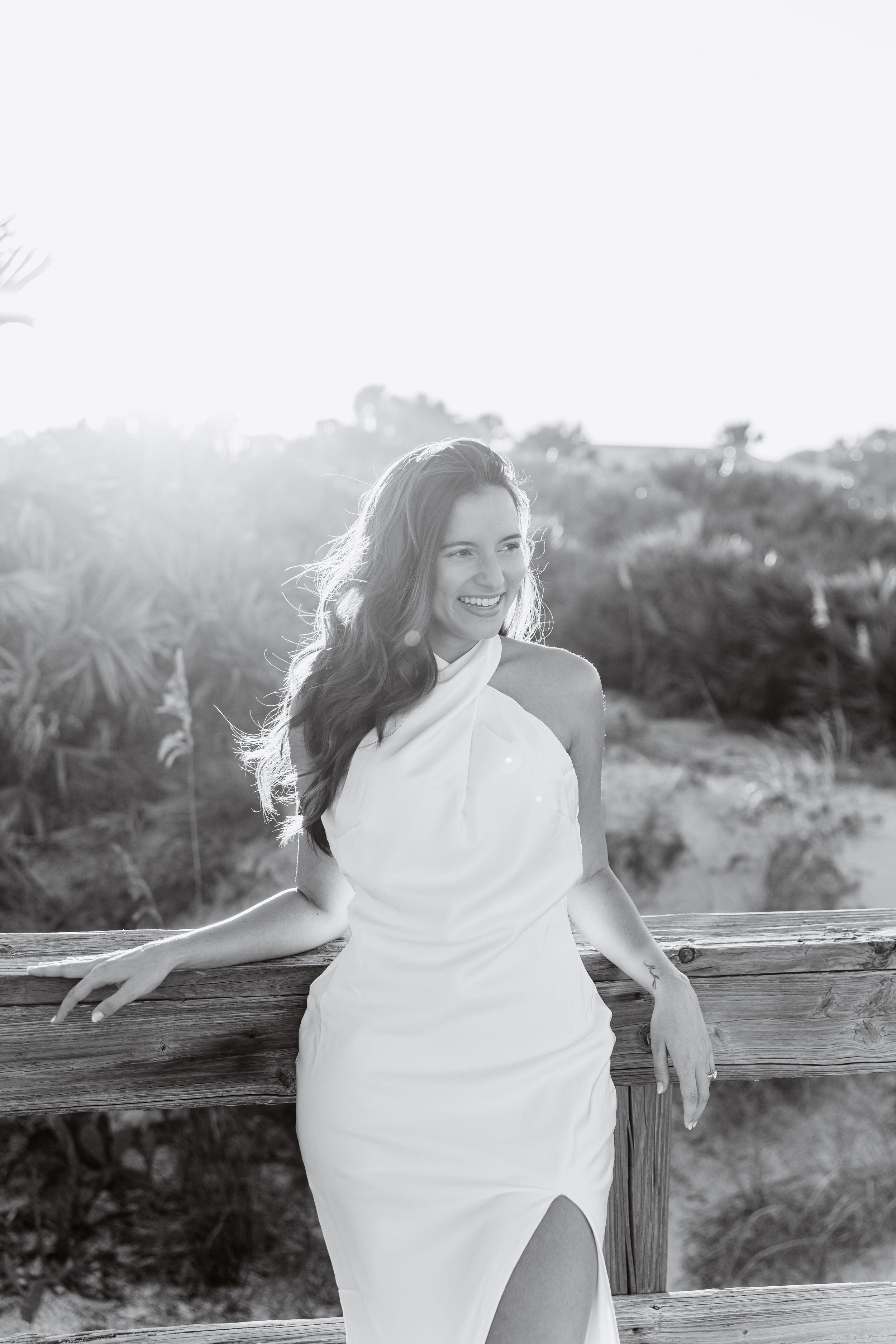 A person in a flowing white dress poses playfully on a wooden fence at sunset in a natural outdoor setting.