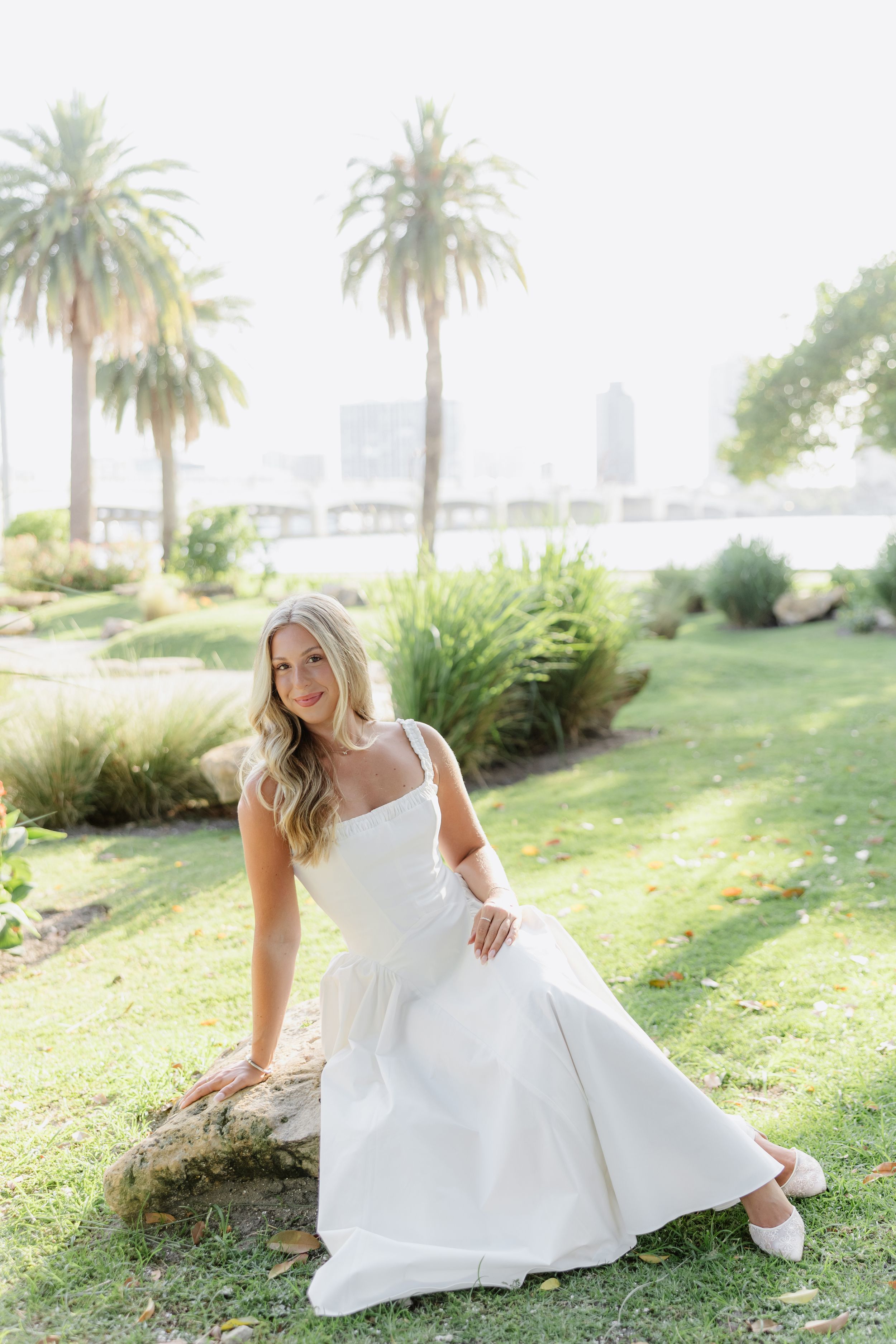 Bride in white gown sits on log in lush garden with palm trees and waterfront skyline backdrop.