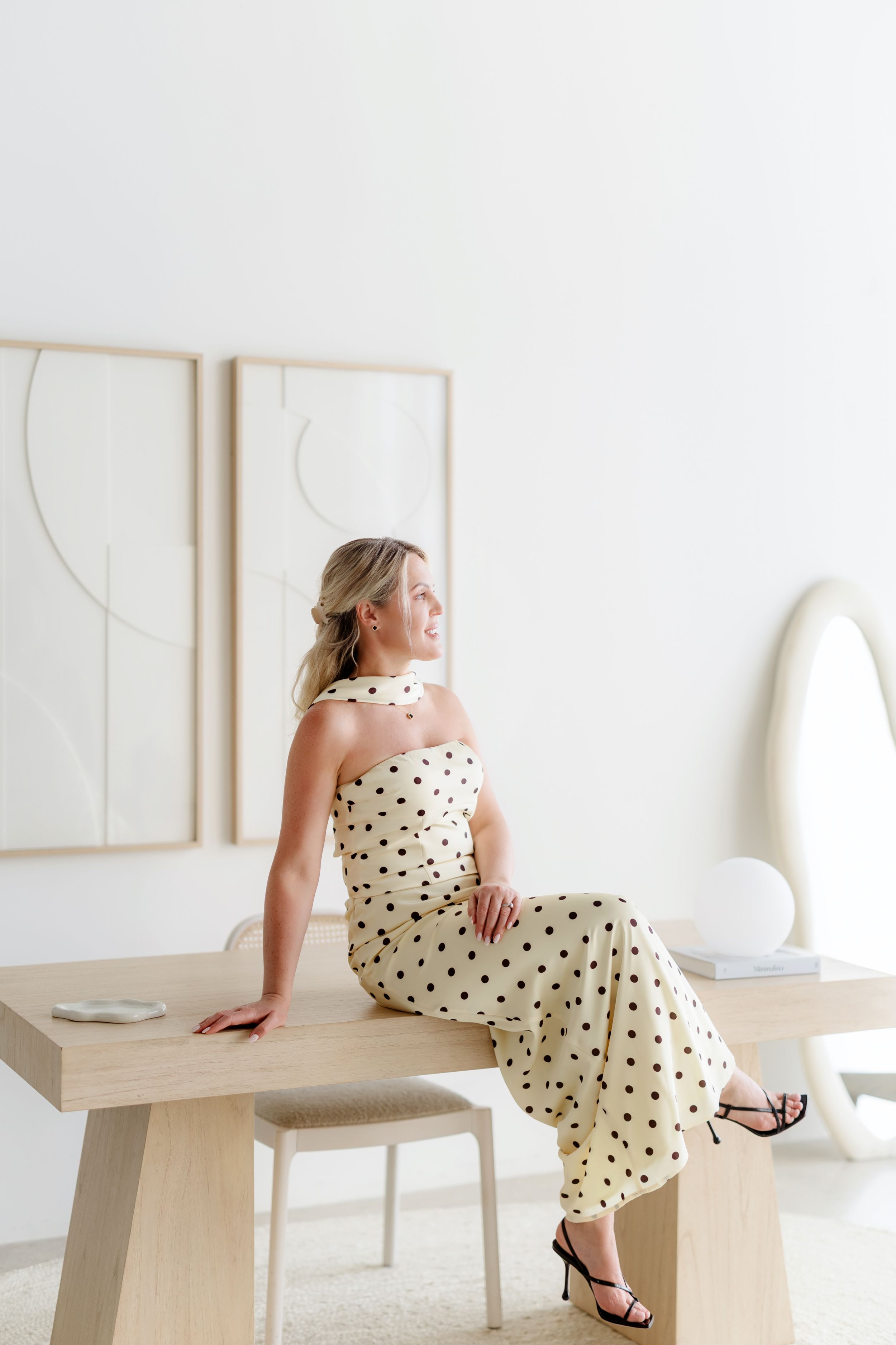 Woman in polka dot halter dress sitting on wooden table in minimalist white room with abstract art.