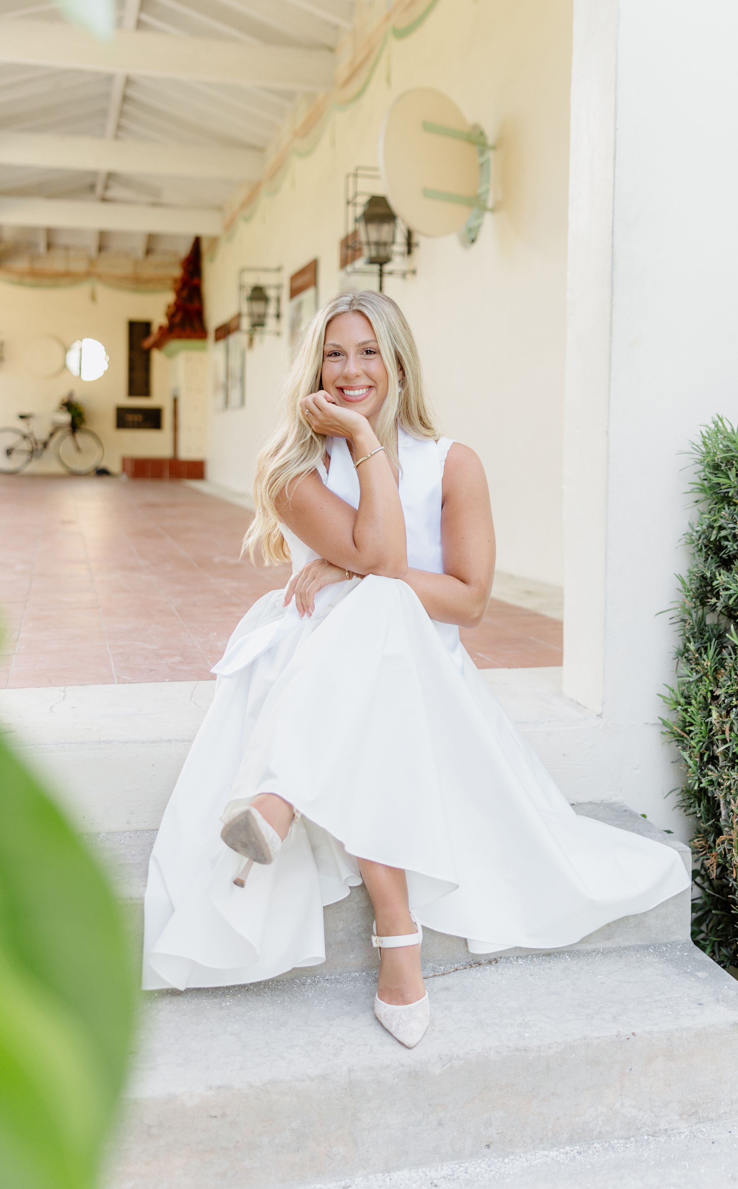 Smiling blonde bride in white sleeveless gown and heels sitting on steps at elegant outdoor venue.