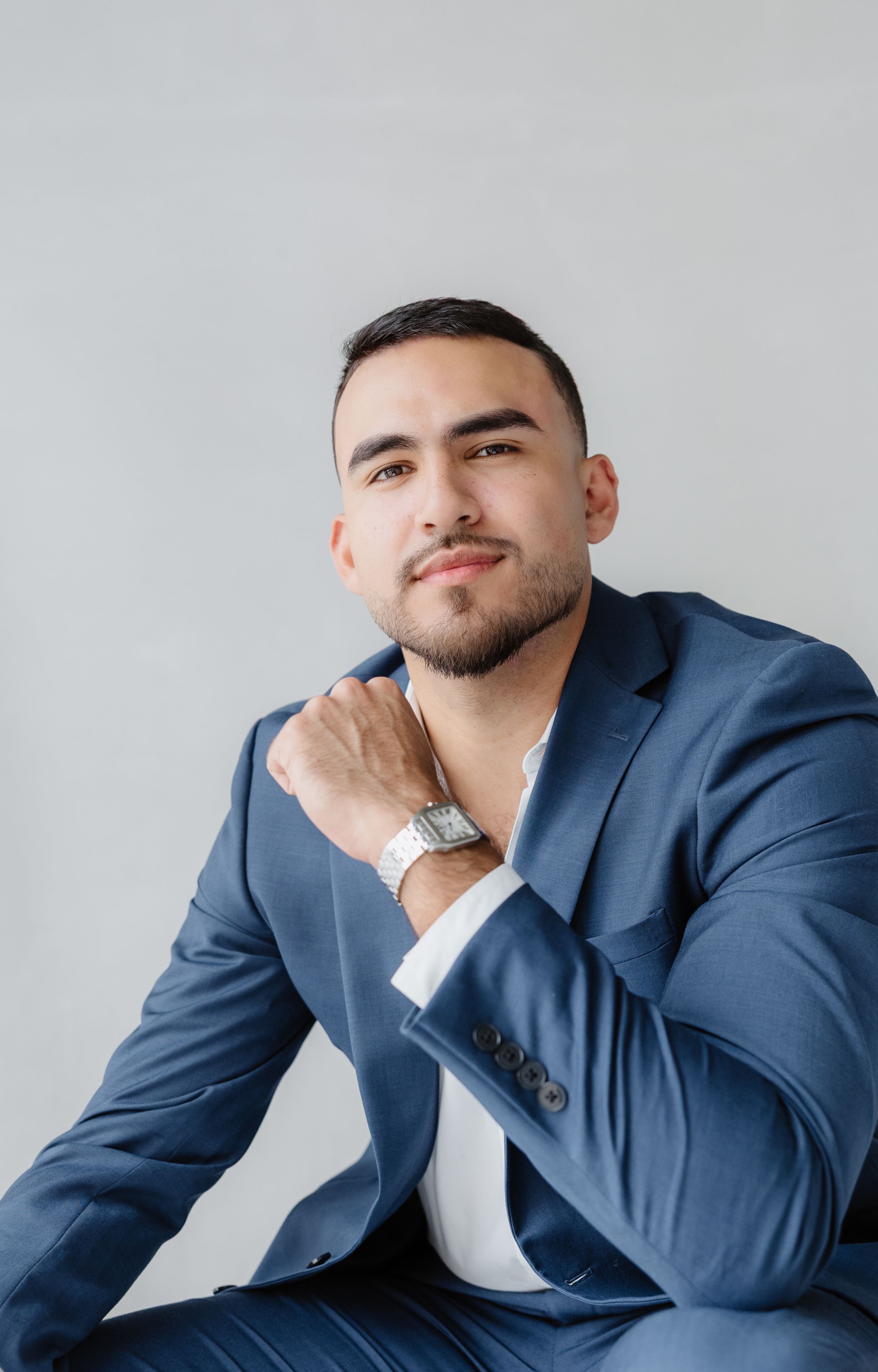 A young man in a blue suit and white shirt sits confidently, wearing a silver watch, against a gray background.