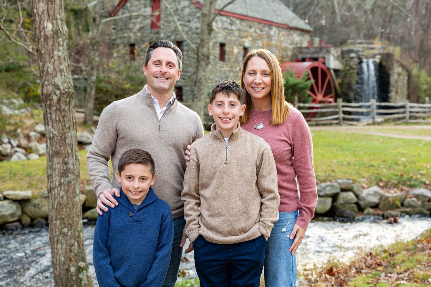 A family group stands poses during a family session in autumn, with colorful fall foliage visible in the background by Lisa West Photography