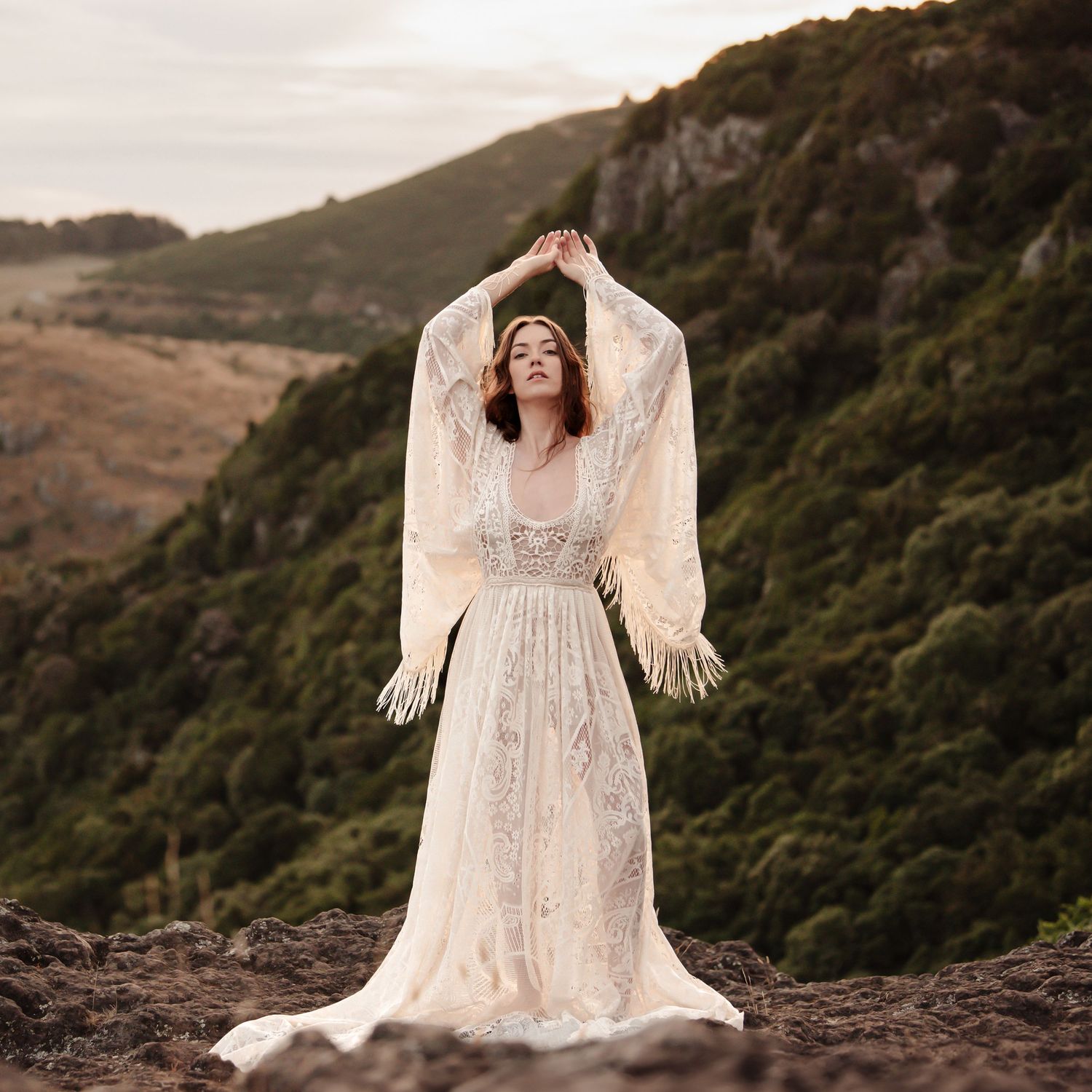 Artistic bridal portrait in vintage lace dress with dramatic sleeves against mountain landscape.