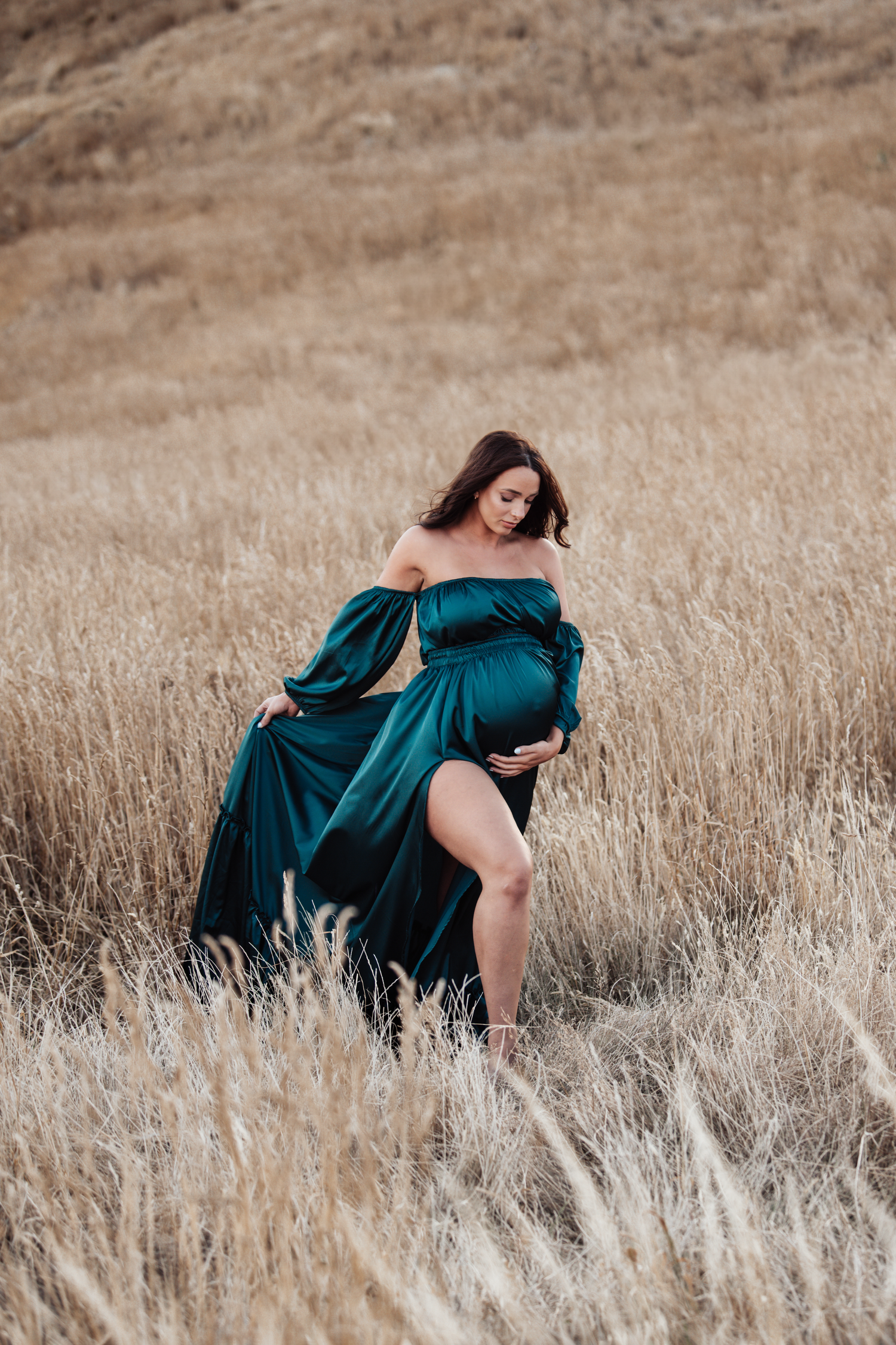 Maternity portrait in teal off-shoulder dress among golden dried grass field.