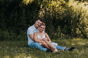 A couple sits together in a sunlit meadow surrounded by wildflowers and tall grass during a romantic outdoor photoshoot.