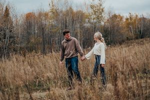 A couple walks together through a scenic autumn landscape with tall dried grass and bare trees in the background.