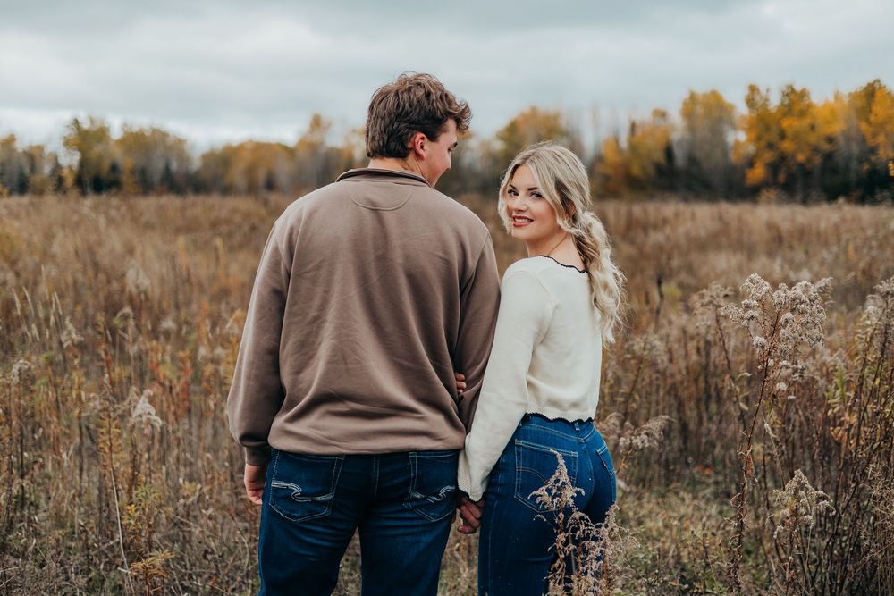 Couple walking together through a golden autumn field during sunset with tall grass and fall foliage in background.