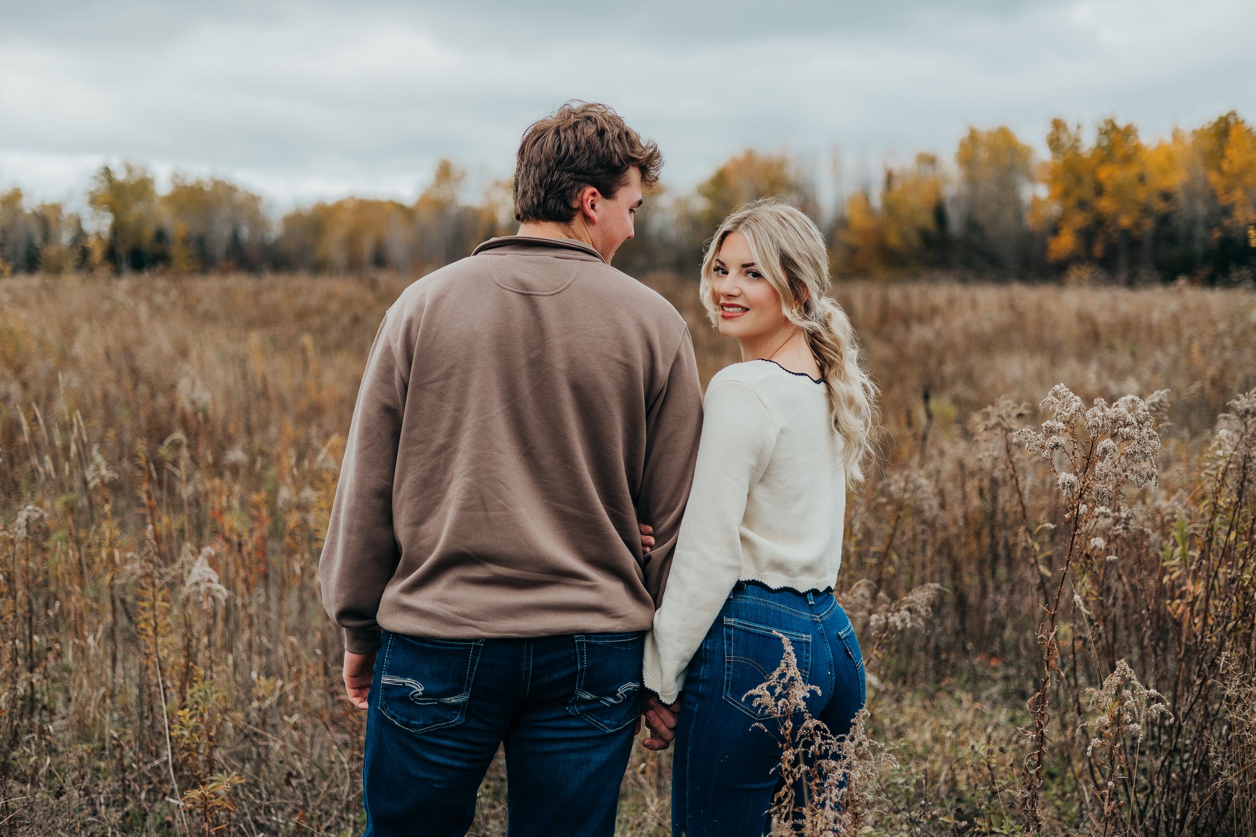 Couple walking together through a golden autumn field during sunset with tall grass and fall foliage in background.