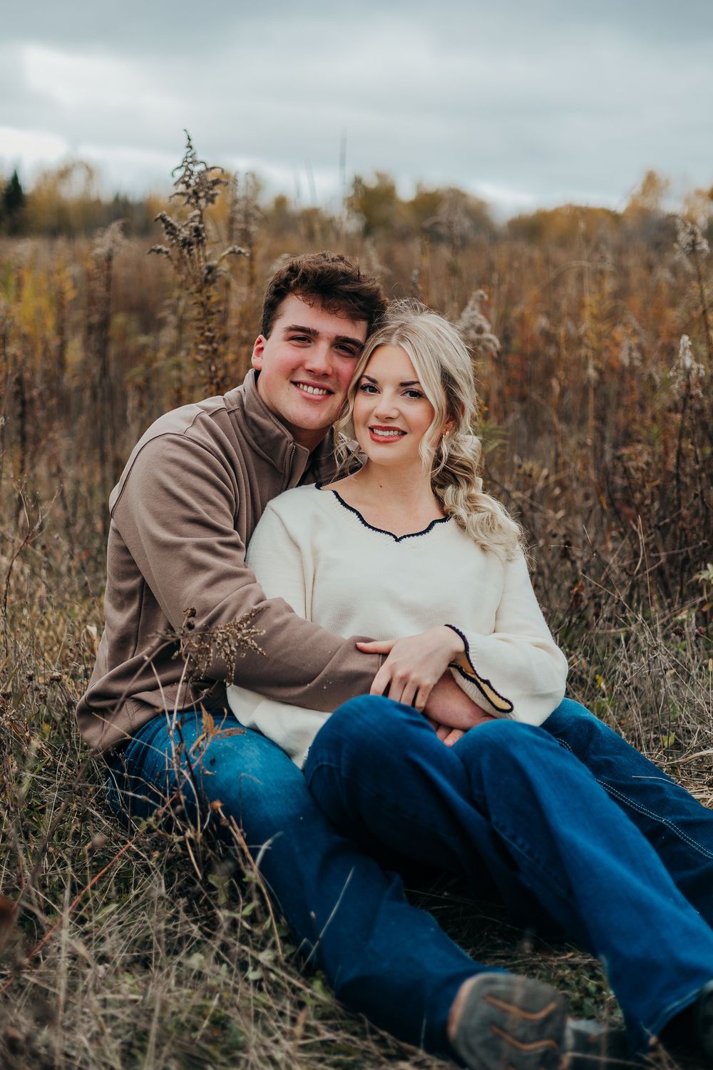 A couple shares a joyful moment together while sitting in a field of tall autumn grasses and wildflowers.