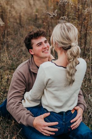 A couple in casual fall outfits share playful candid moments together in a field with autumn foliage in the background.