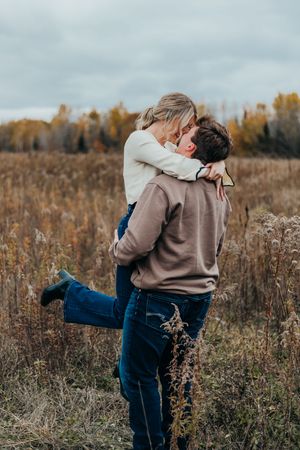 A couple embraces playfully in a rustic outdoor setting surrounded by fall foliage and dried grasses.