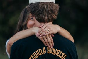 A person in a black t-shirt with tequila sunrise text embraces someone while standing outdoors.