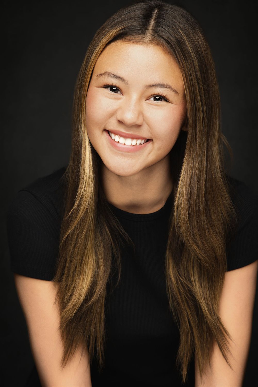 Actor headshot of young girl with long brown hair wearing black top against dark background. Shot by Mika Larson Photography.