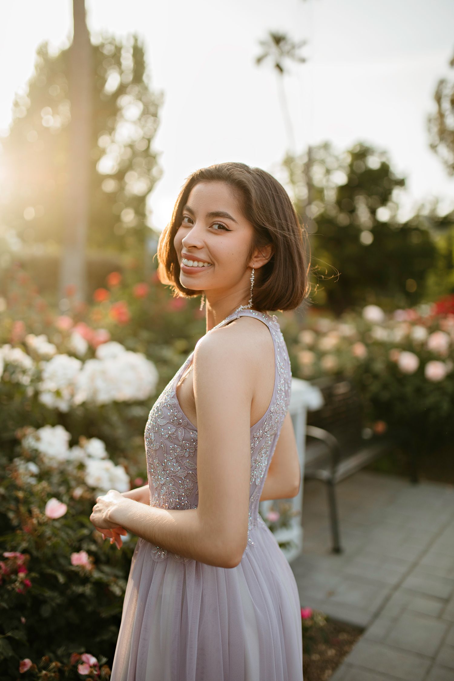 Sacramento Graduate in a lavender dress poses in a rose garden with blooming roses during golden hour sunset.