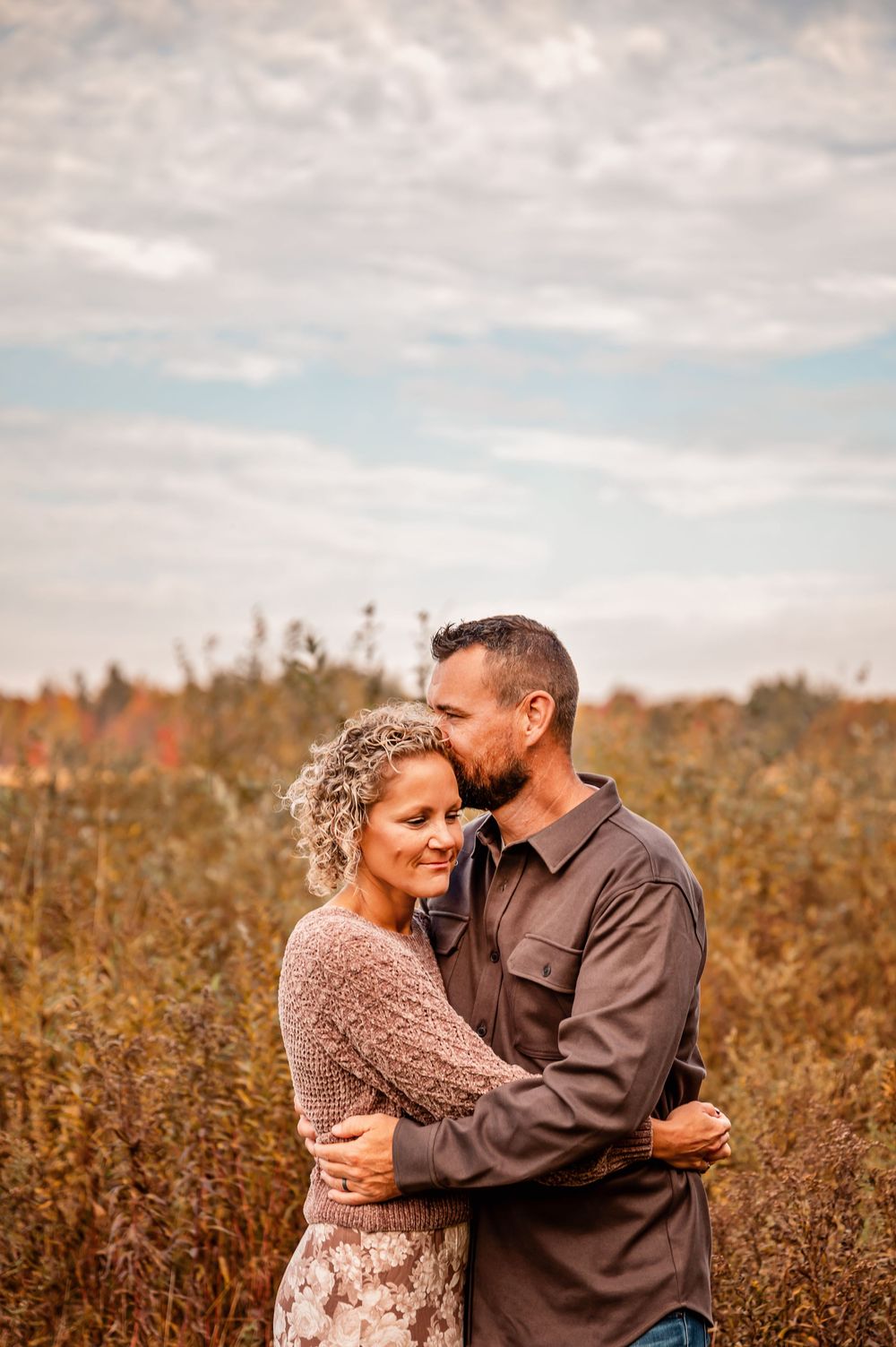A couple embraces lovingly in a golden autumn field under cloudy skies.