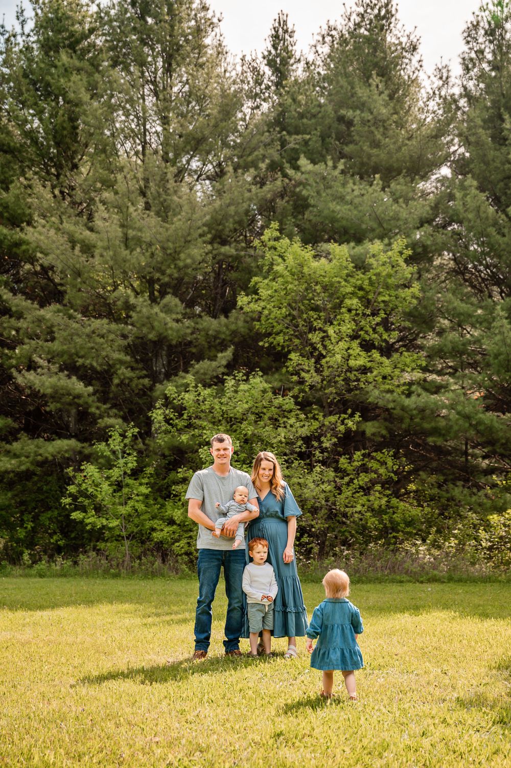 Family poses together in grassy field with tall pine trees in background.