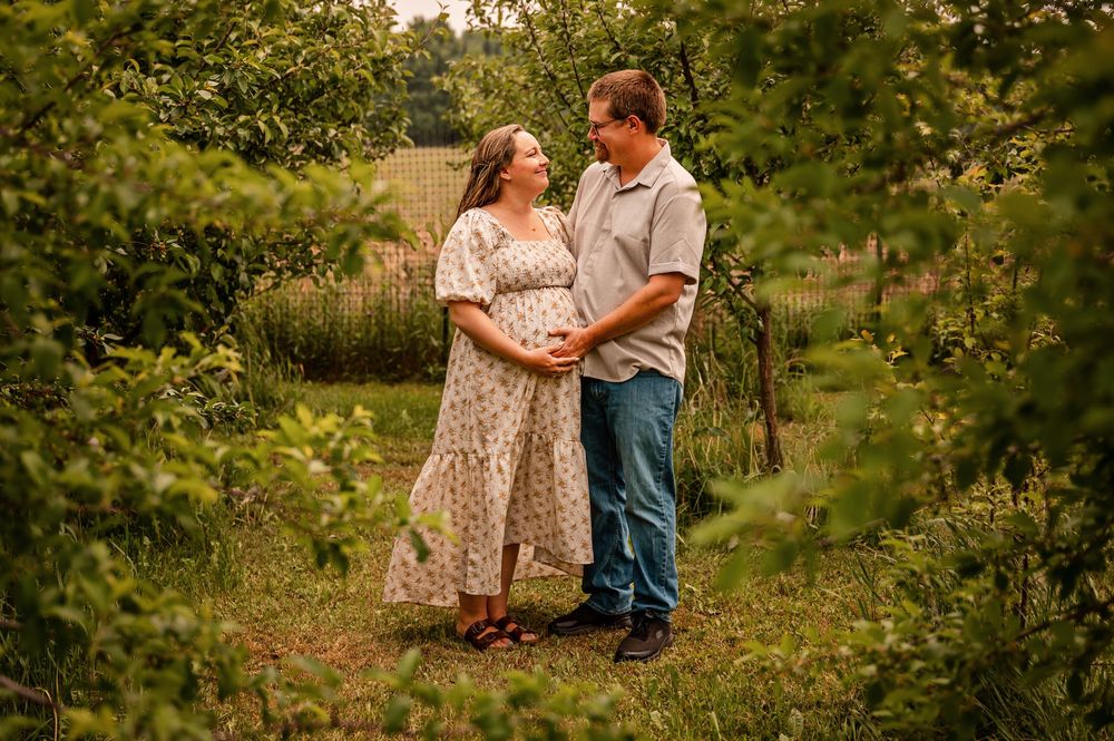 A couple stands together in a sunlit orchard during a maternity photo session, surrounded by green foliage.