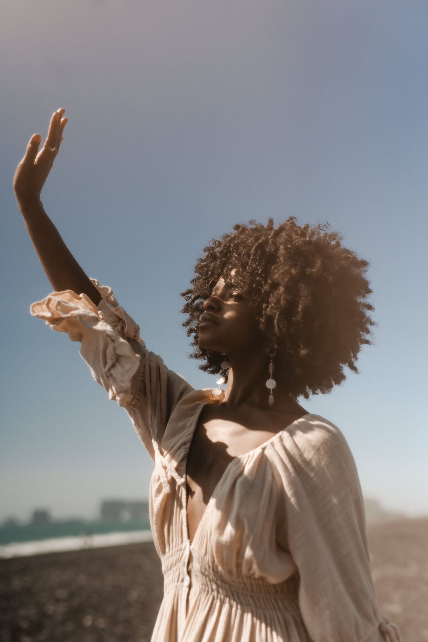 Person in flowy white dress reaching toward sky with natural curly hair.