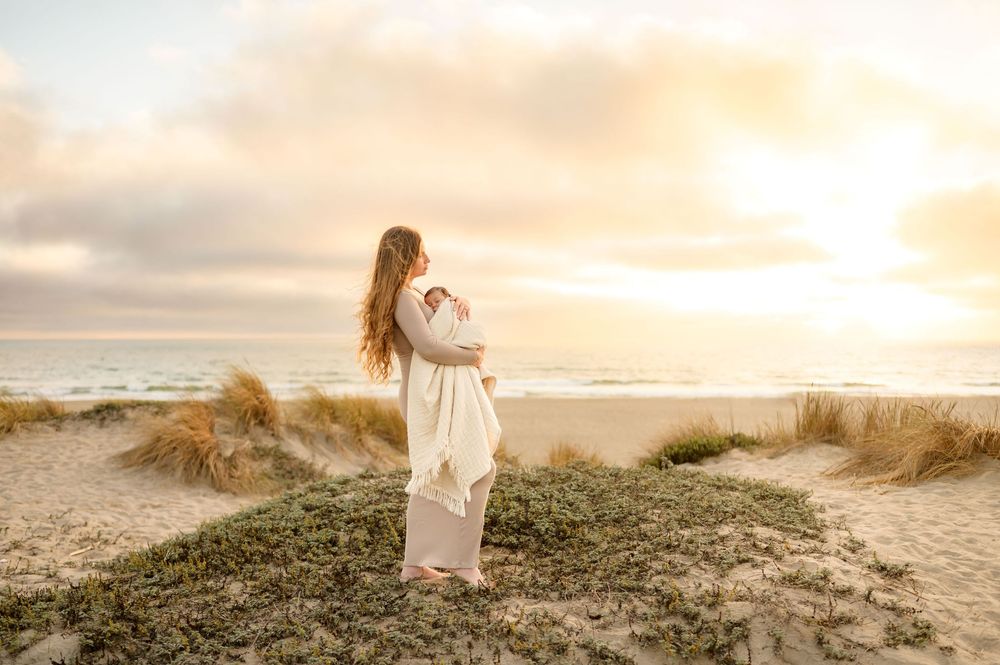 woman stands on beach dunes during beach newborn photoshoot at sunset by Ventura photographer