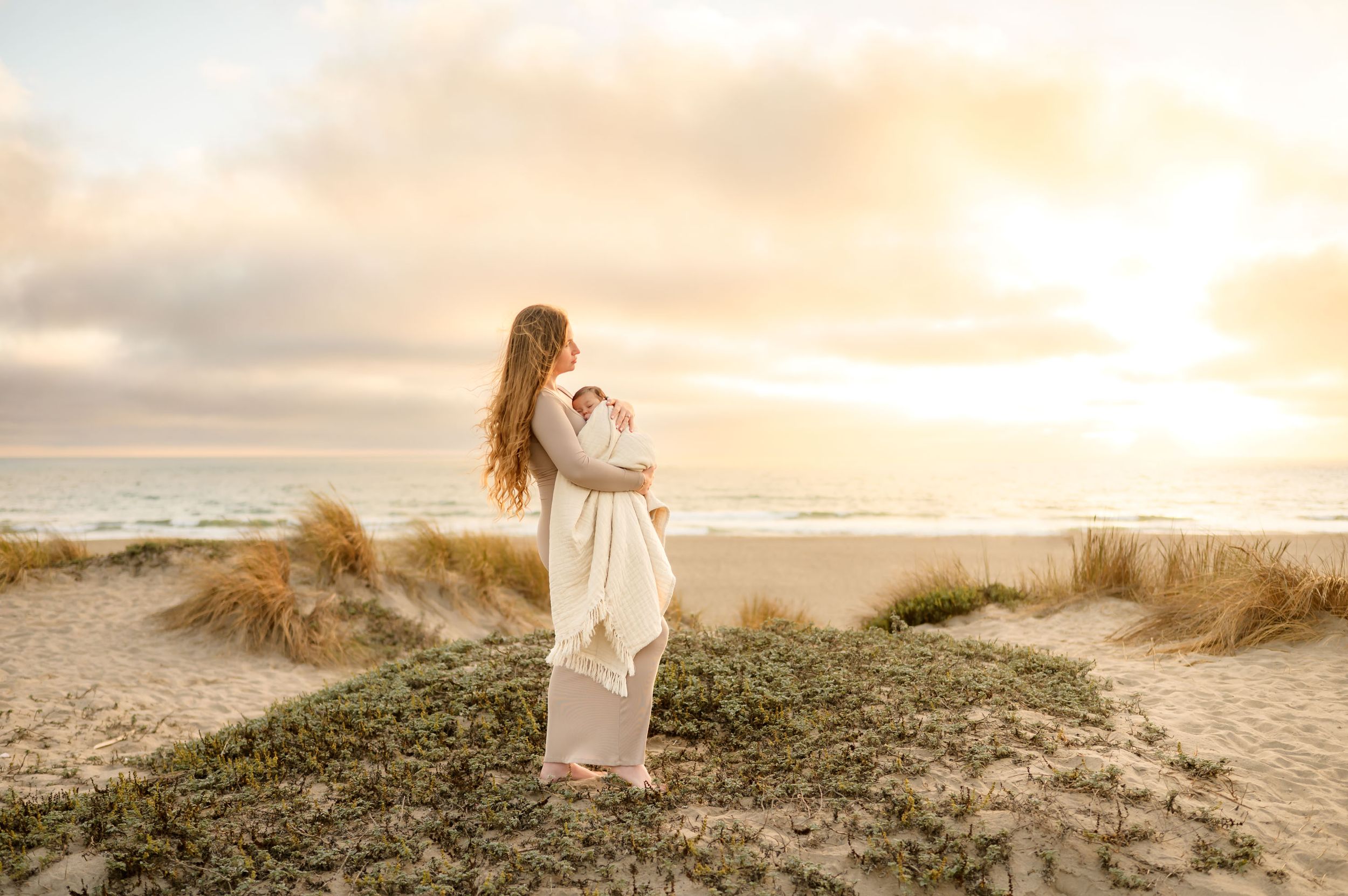 woman stands on beach dunes during beach newborn photoshoot at sunset by Ventura photographer
