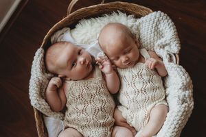 Two sleeping babies wearing matching cream knit outfits lie together in a cozy woven basket with textured blankets.