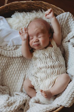 Sleeping infant wearing white knit dress lying on cream colored blanket in soft natural light.
