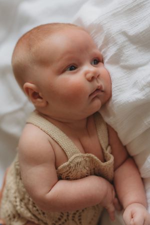 A newborn baby wearing a tan knit romper lies peacefully on white bedding in a series of close-up photos.