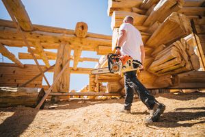 Worker carrying a chainsaw at a log cabin construction site with stacked wooden logs under a clear blue sky.