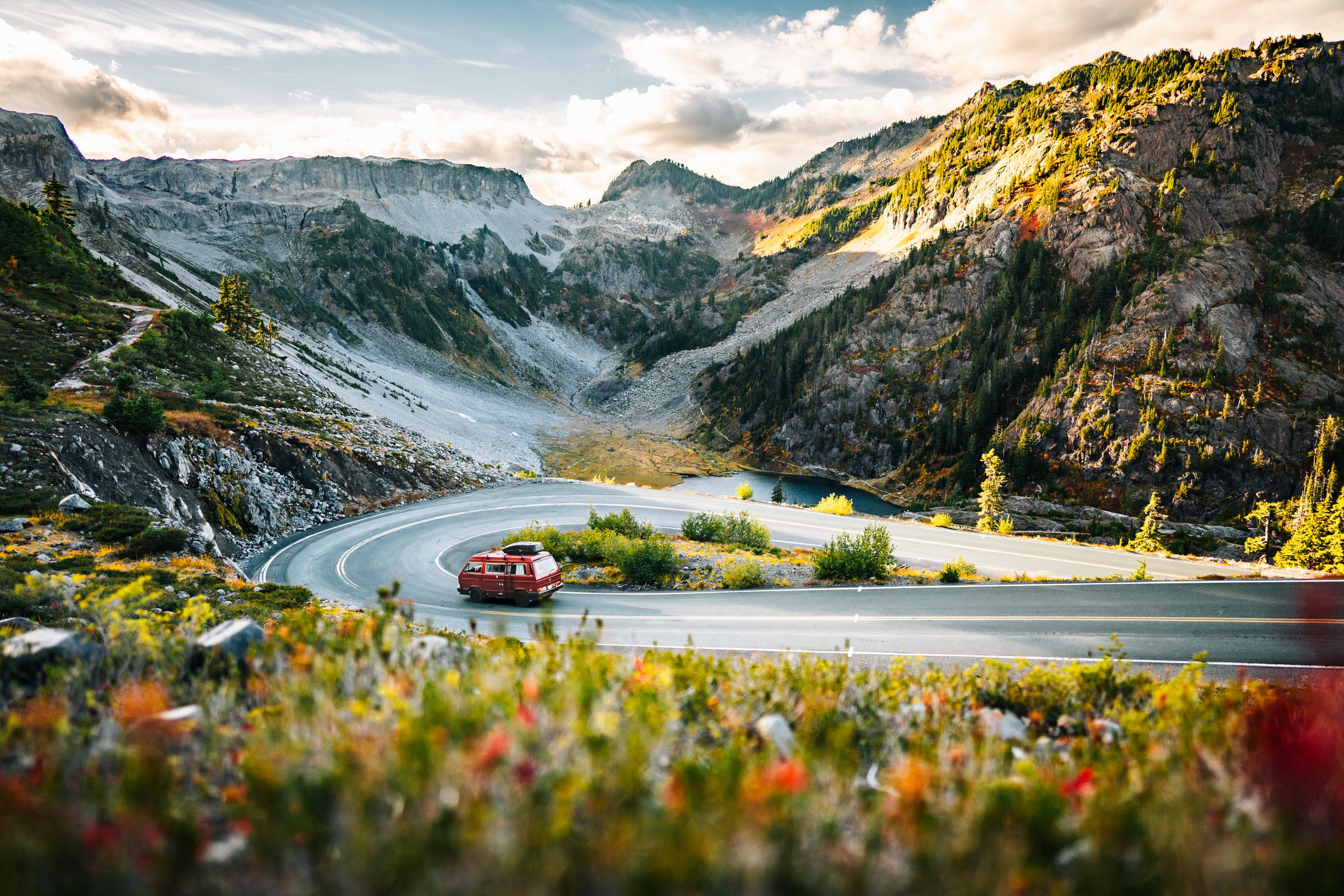 Red car navigating a winding mountain road surrounded by colorful wildflowers, rocky peaks, and a serene alpine lake.