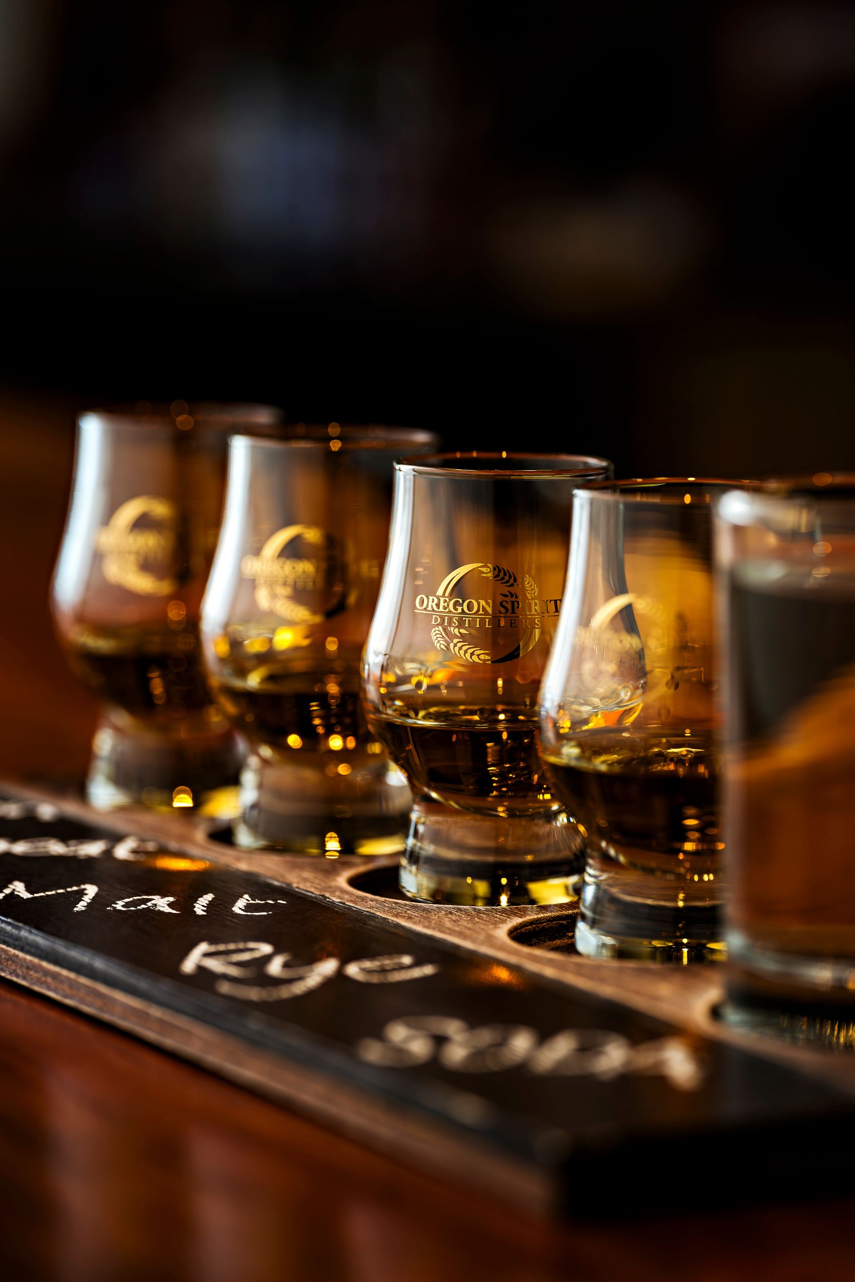 Several whiskey glasses with branded logos sit on a dark wooden tasting board in warm ambient lighting.