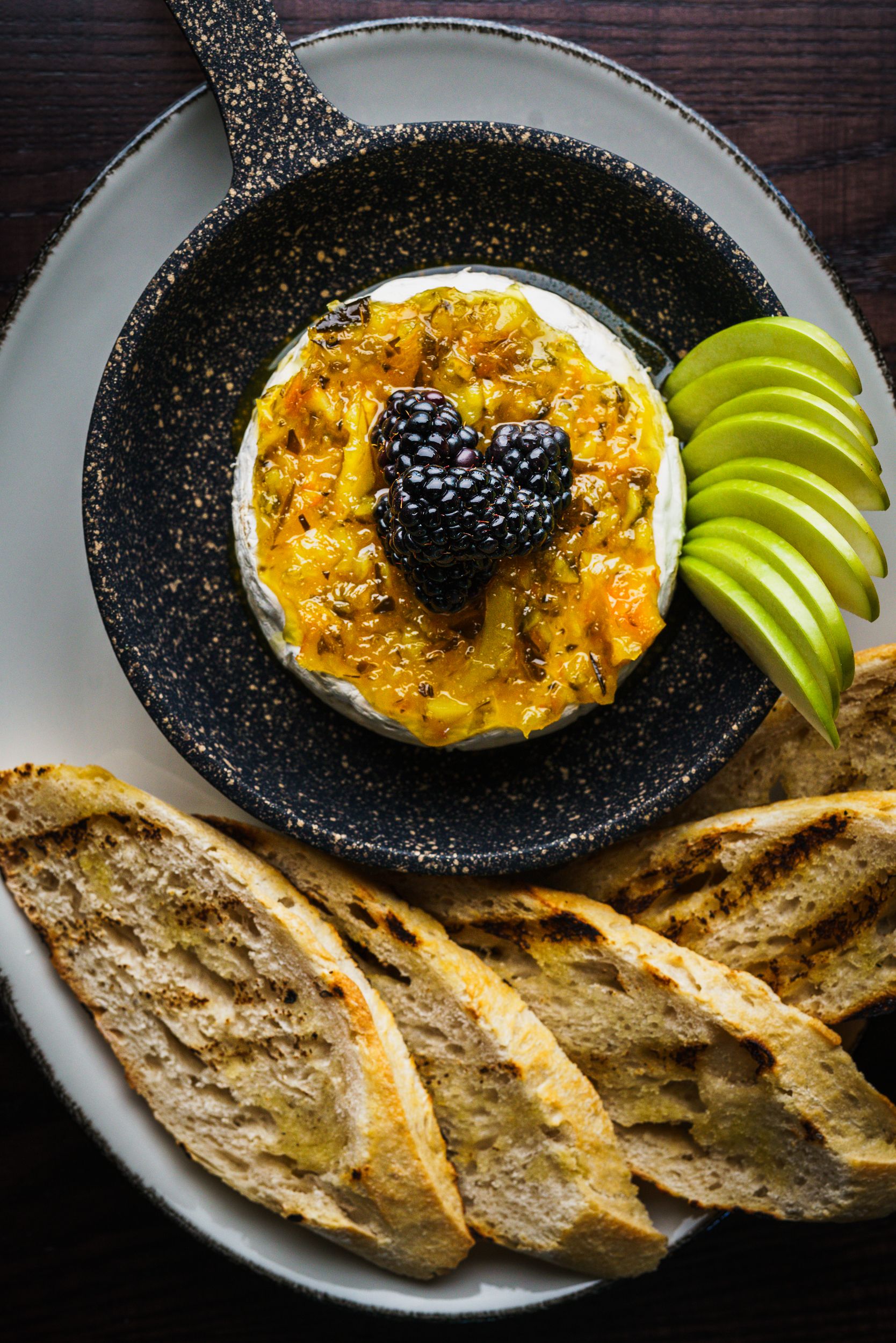 Gourmet cheese topped with blackberries and apple slices served with toasted bread served in a skillet.