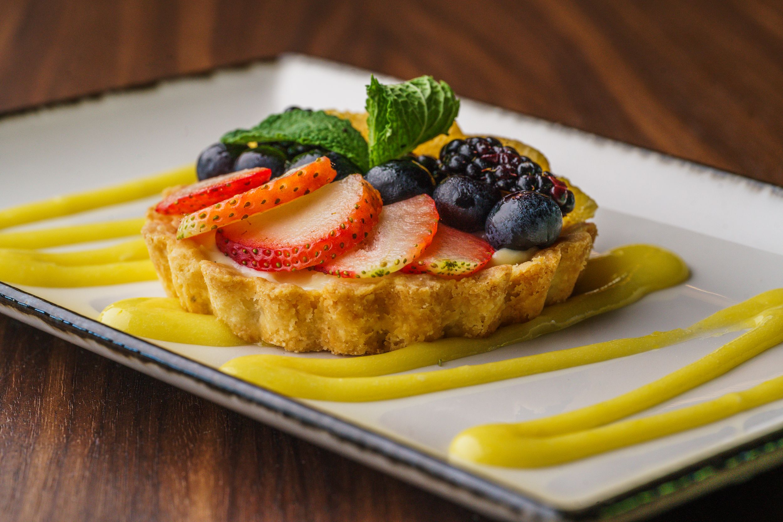 Fresh fruit tart with strawberries and blueberries on white plate with yellow sauce drizzle.