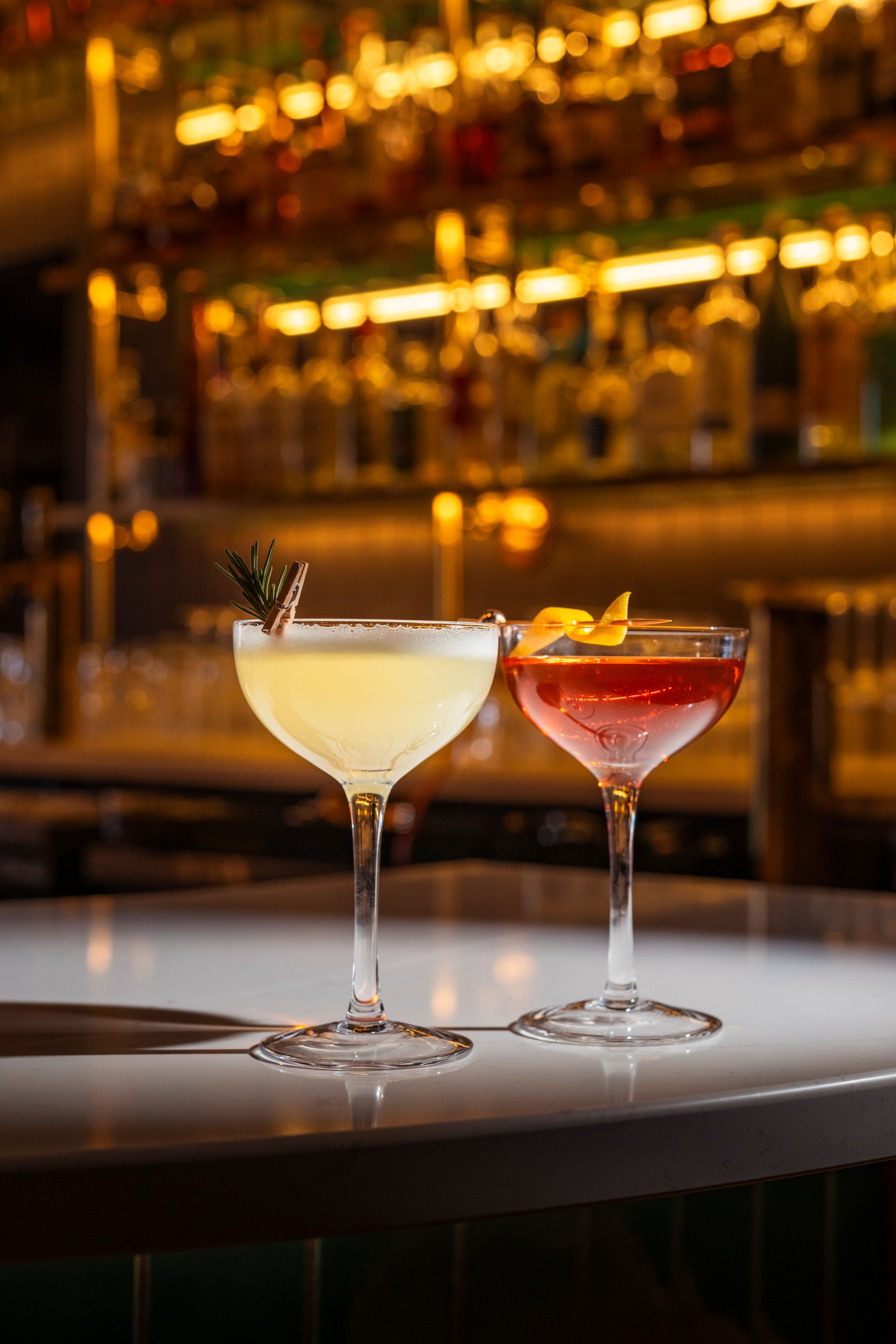 Two elegant cocktails on a bar counter with blurred shelves of liquor bottles illuminated in warm lighting in the background.