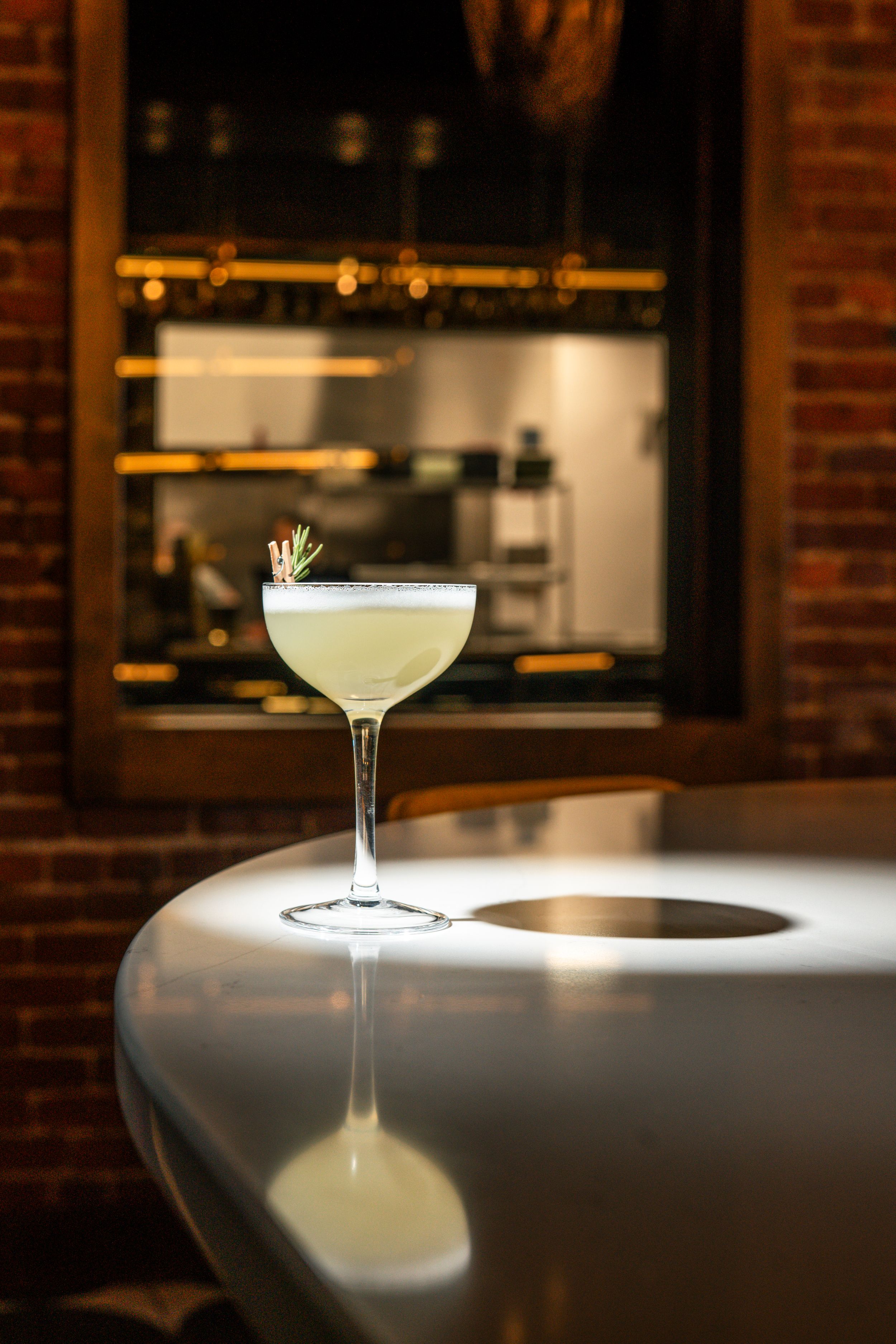 Elegant craft cocktail with frothy top served in coupe glass on sleek bar counter with moody lighting and exposed brick wall.
