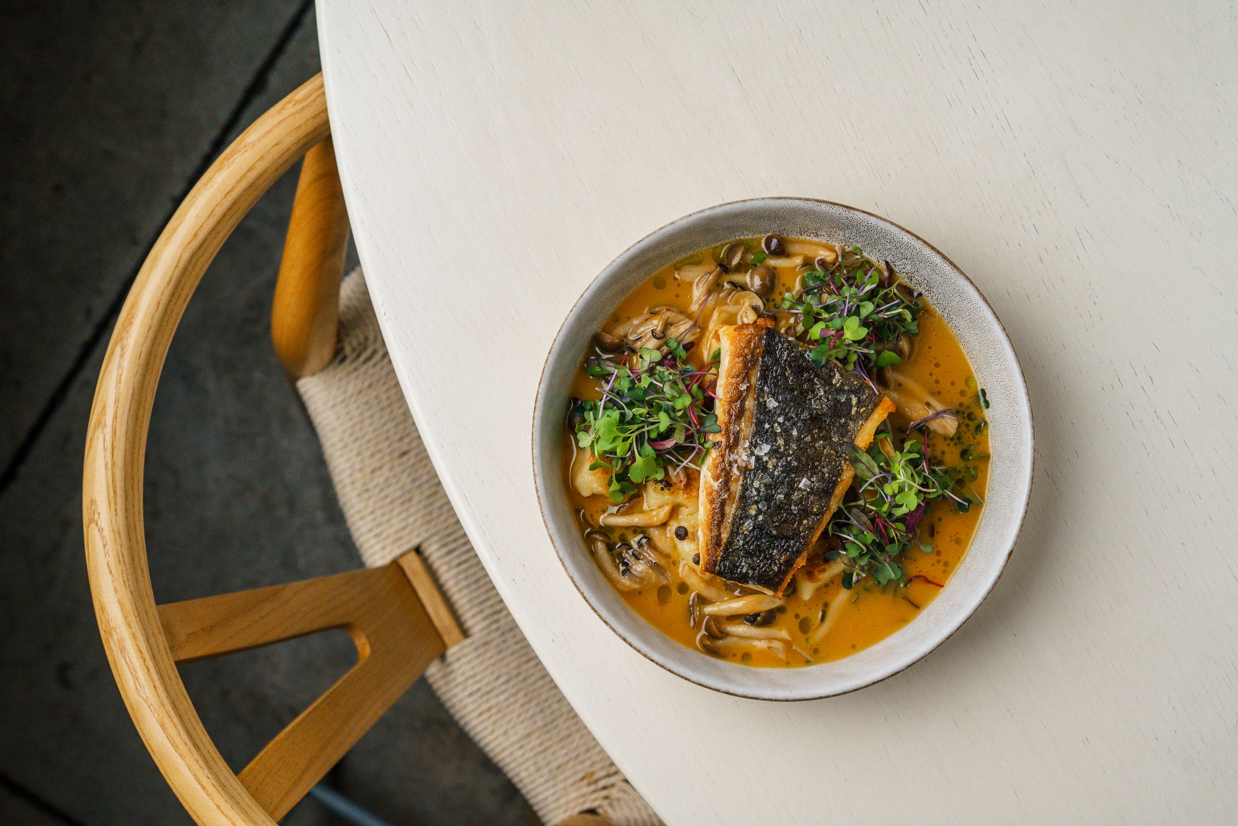 Grilled mackerel fillet served in a white bowl with aromatic broth and fresh herbs on a white surface.