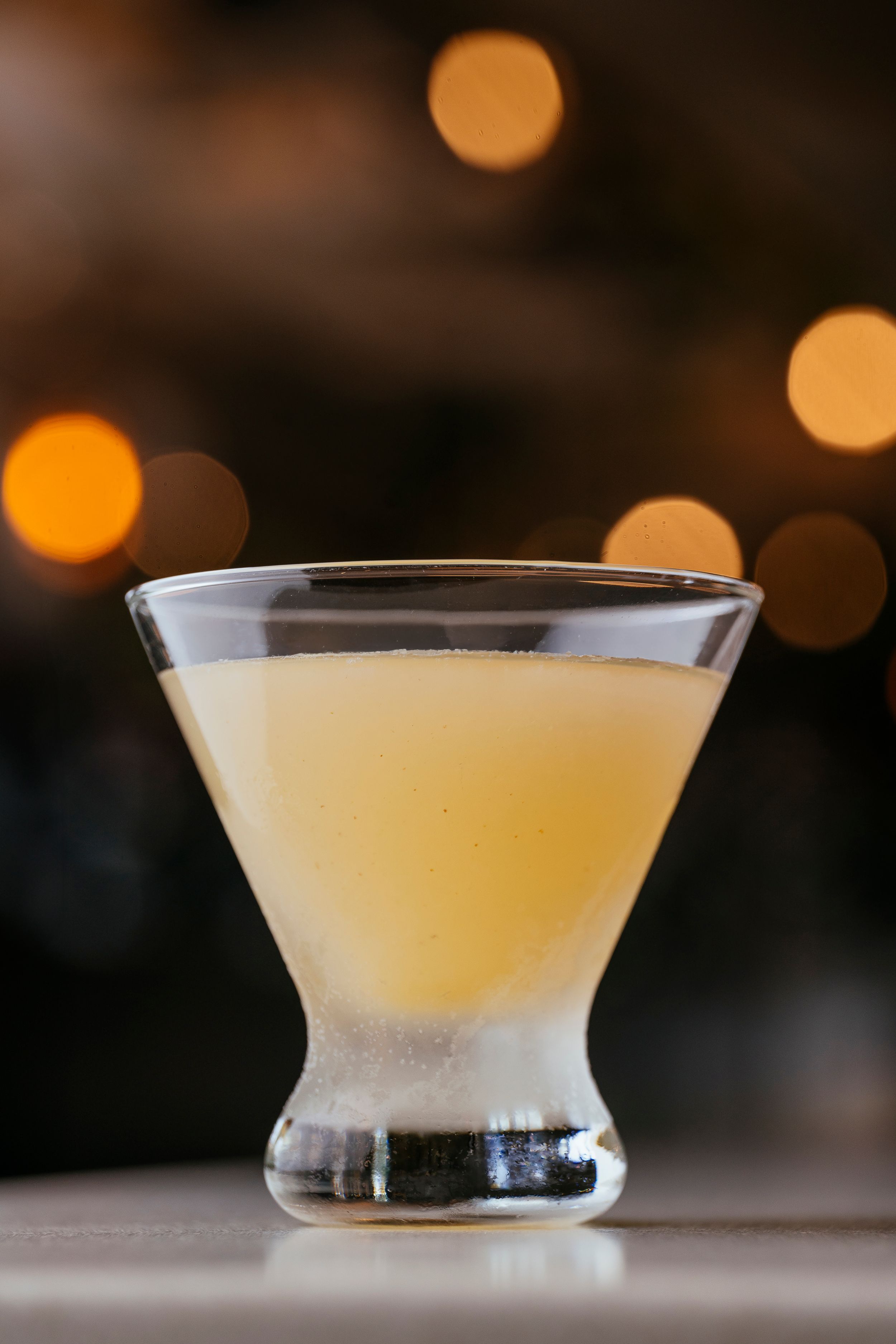 A yellow cocktail served in a martini glass with blurred warm lights in the background creating a bokeh effect.