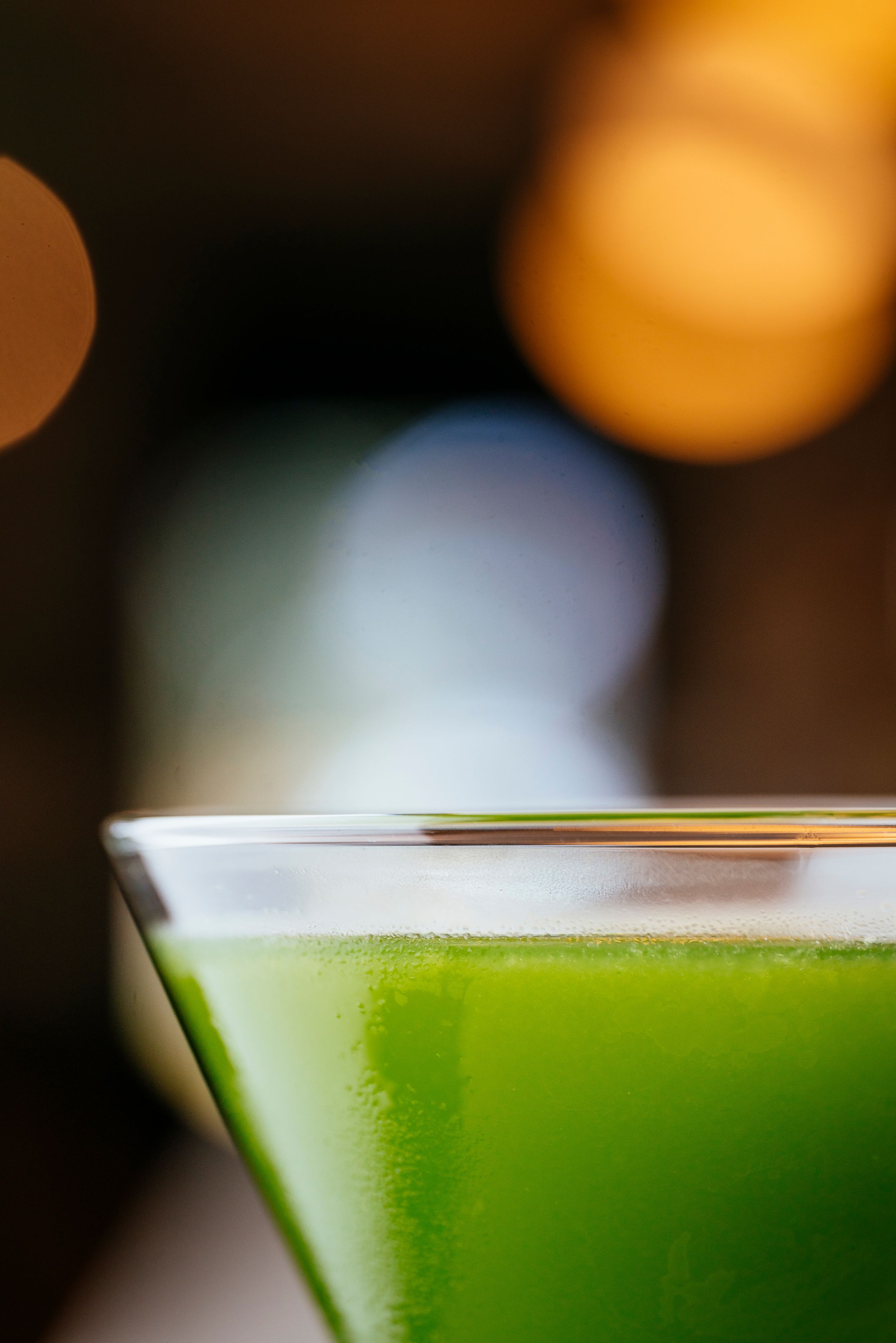 Close-up view of a bright green cocktail in a martini glass with blurred bokeh lights in the background.
