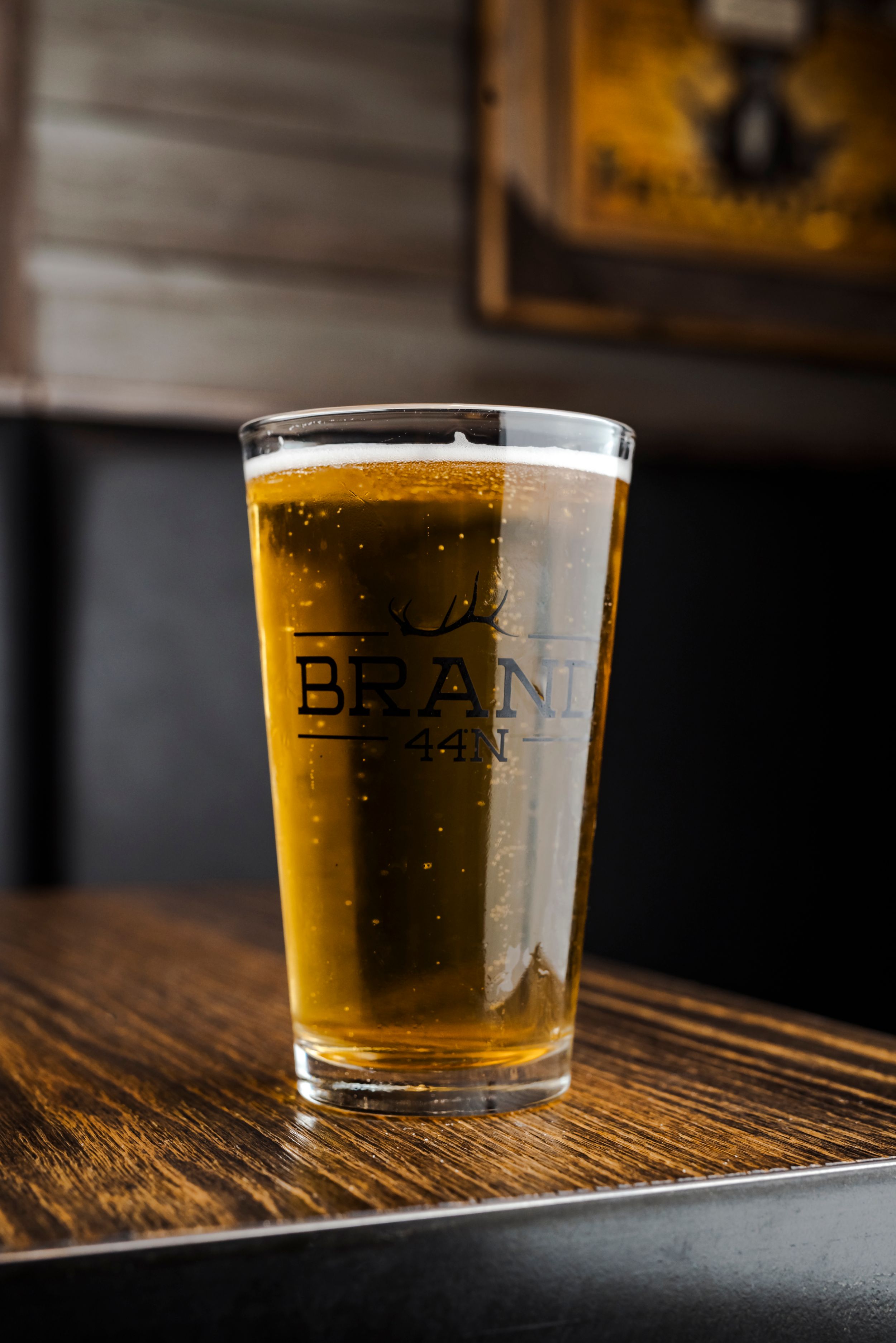 A pint glass of golden beer with the word 'BEANI' etched on it sits on a dark wooden table in dim lighting.