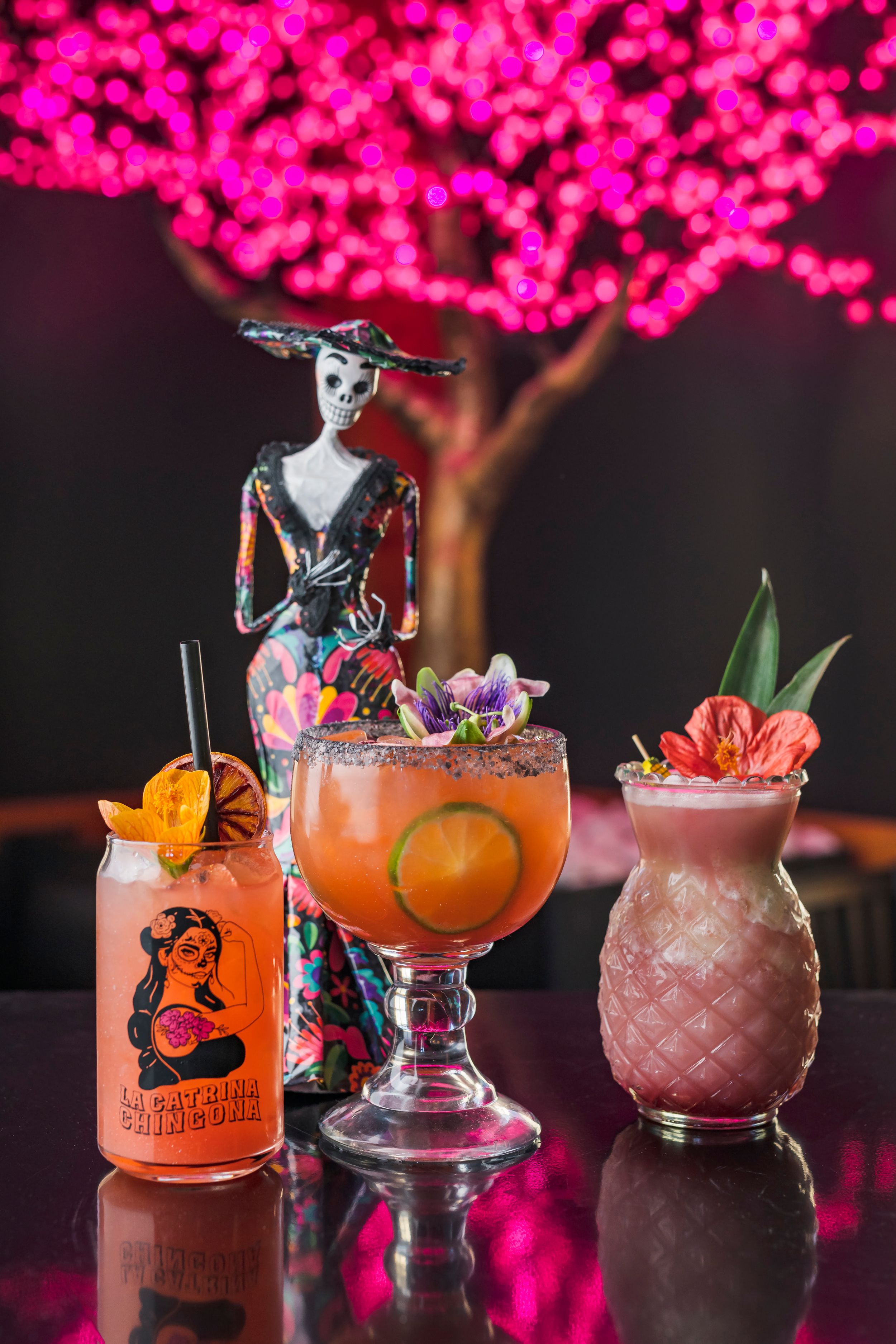 Three tropical cocktails with colorful garnishes displayed under a pink-lit tree with decorative figure topper.
