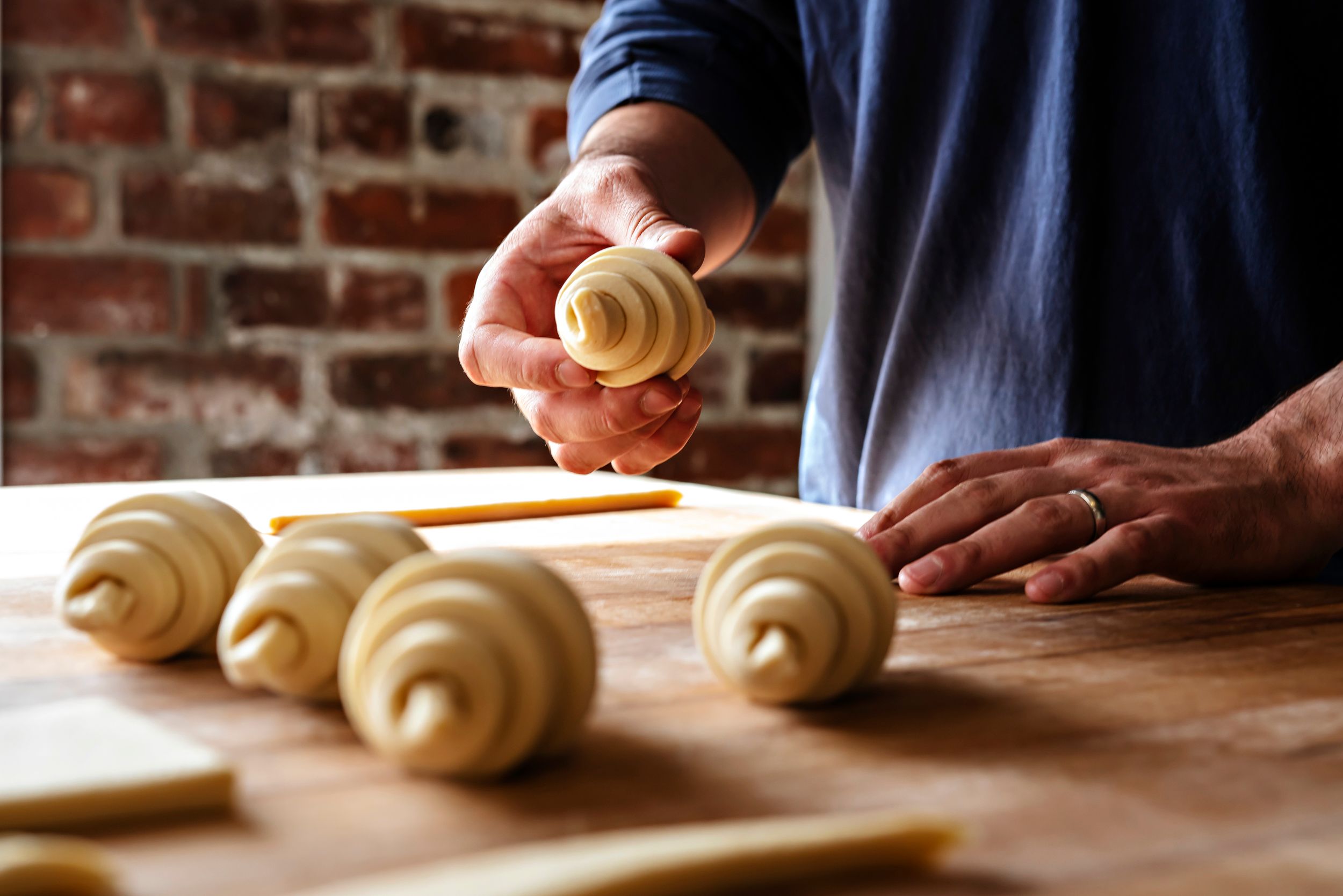 Hands rolling fresh croissant dough on a wooden surface with a brick wall background.