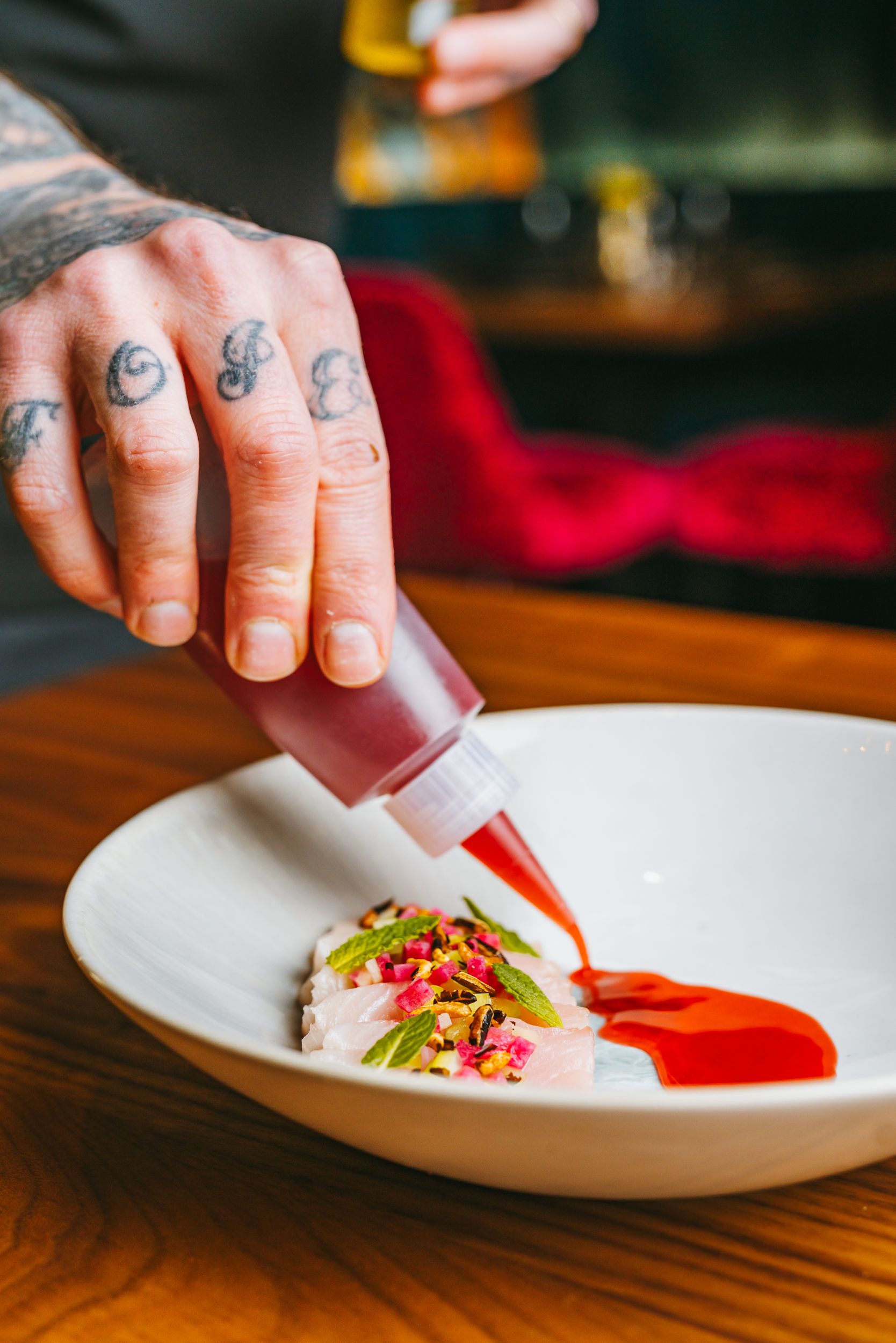 Chef squeezing red sauce onto a plated dish with tattooed hands.